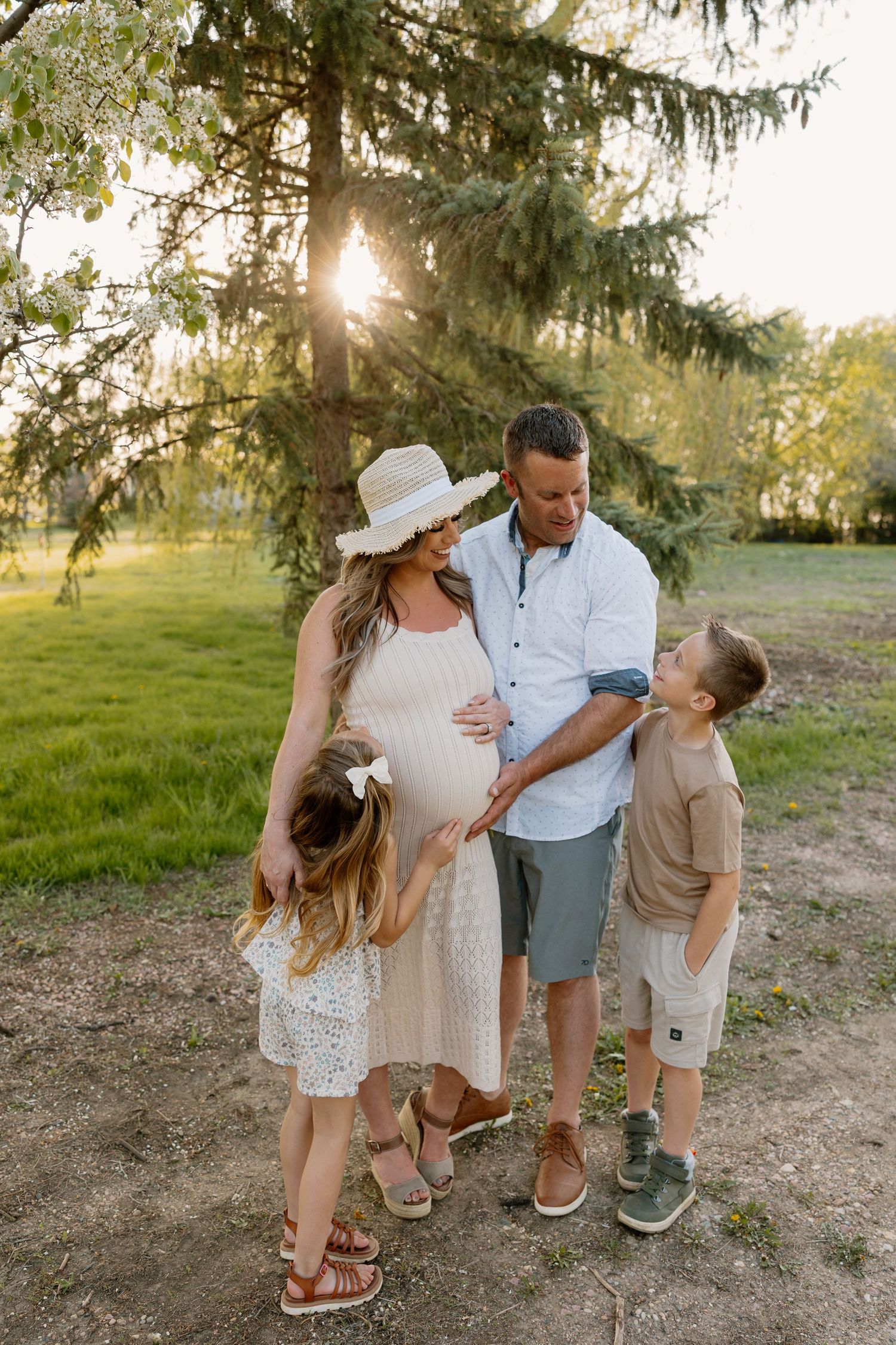 A family enjoys a tender moment together outdoors on a sunny evening beneath towering evergreen trees.