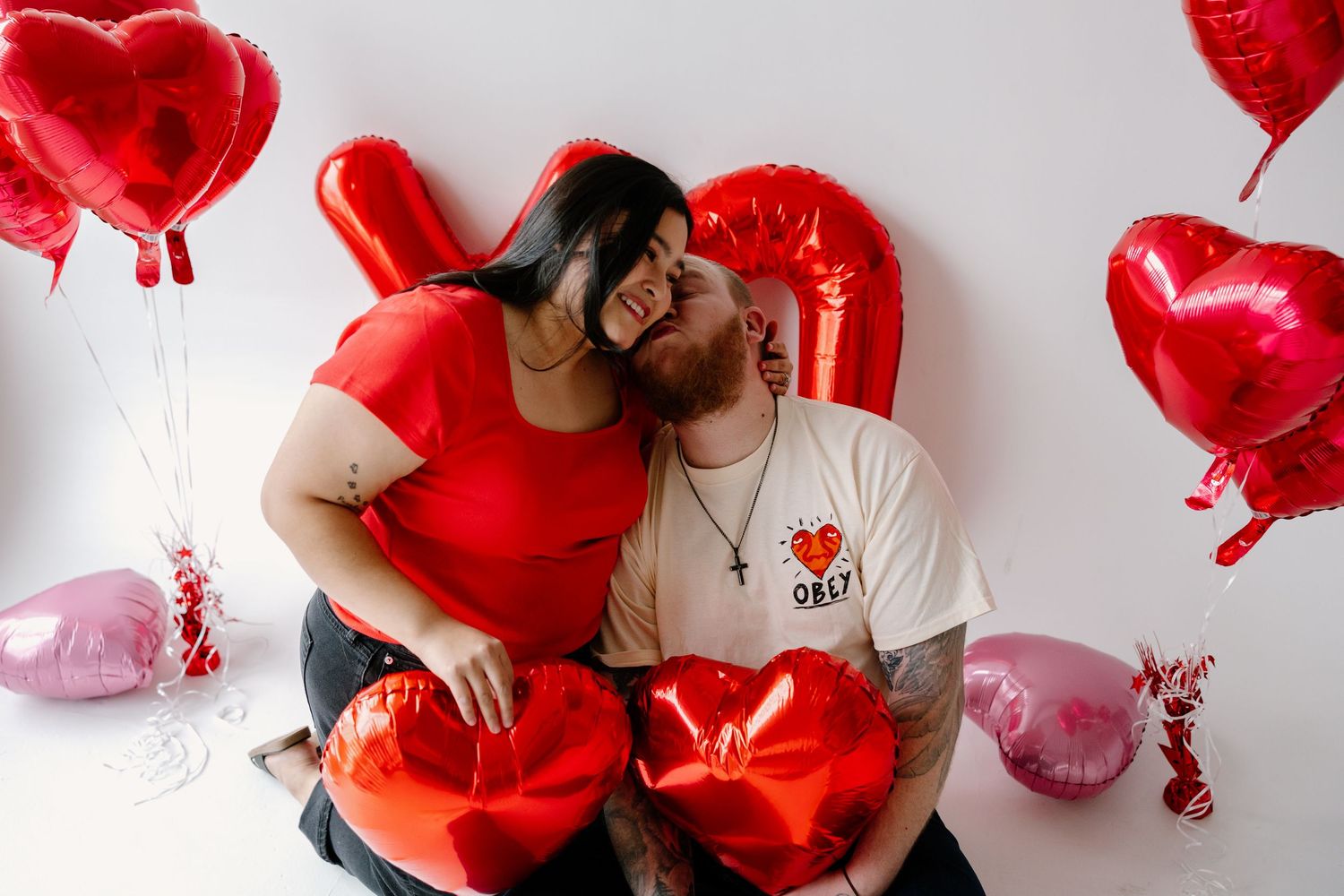 Romantic couple poses with red and pink heart-shaped balloons against white background on Valentine's Day.