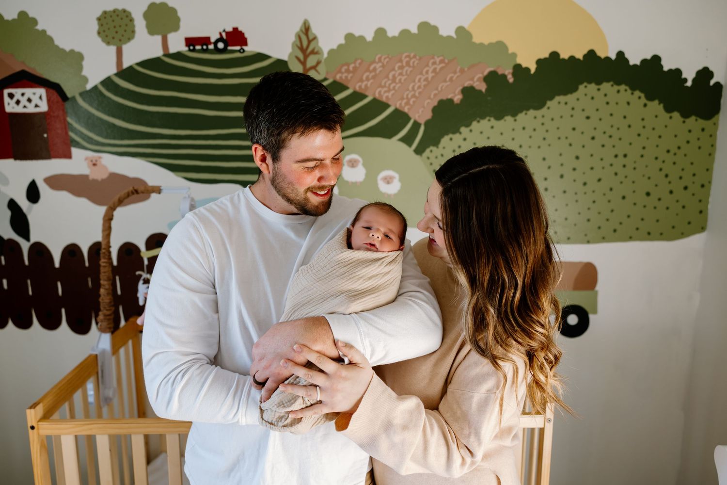 New parents smiling and holding their newborn baby in a cozy nursery with a colorful farm mural on the wall.