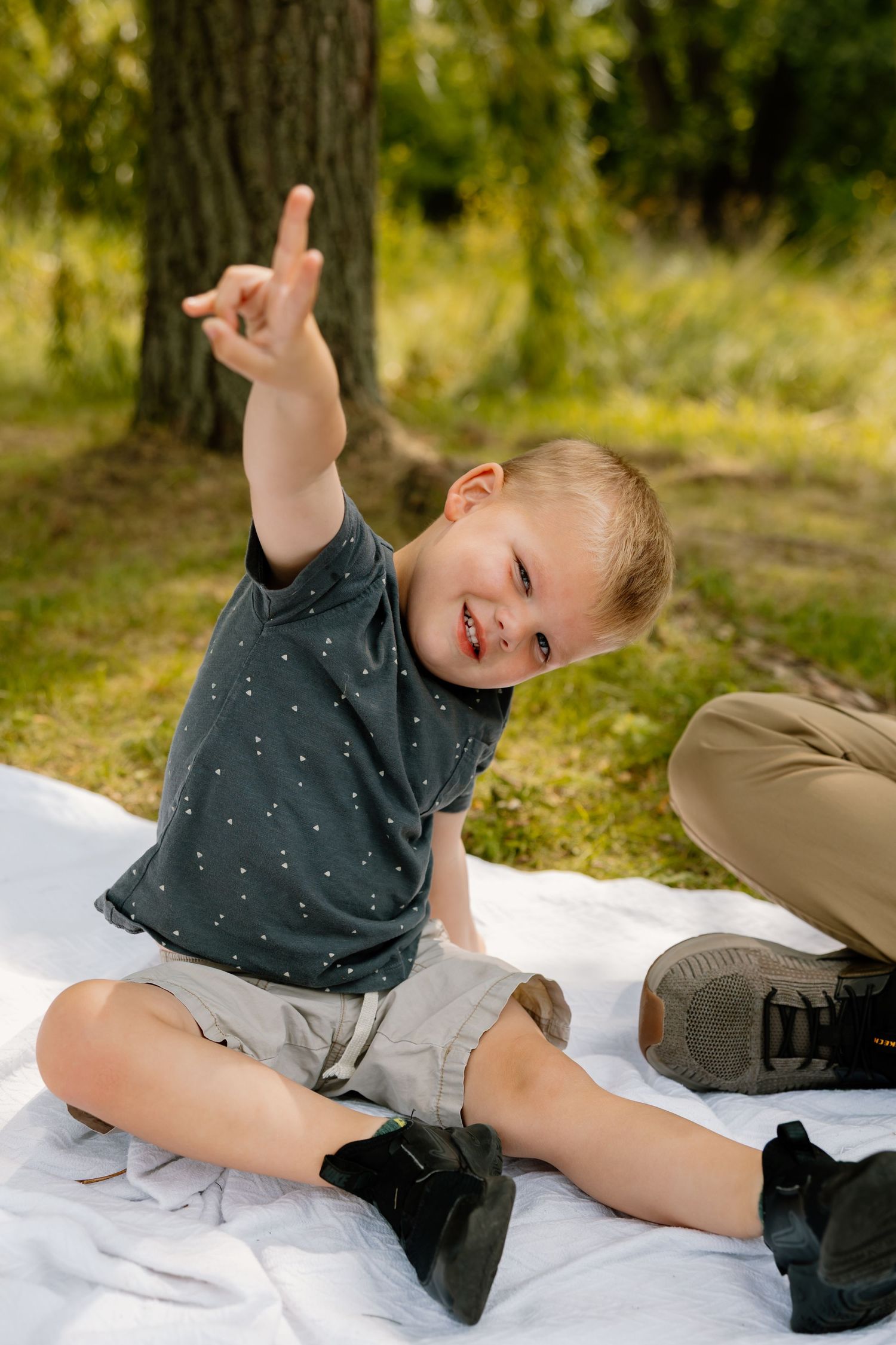 Child in gray shirt sits on white blanket under tree making playful gesture.