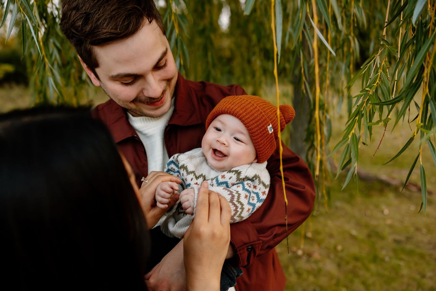 Happy baby in orange knit hat laughing with parents under willow tree in autumn park.