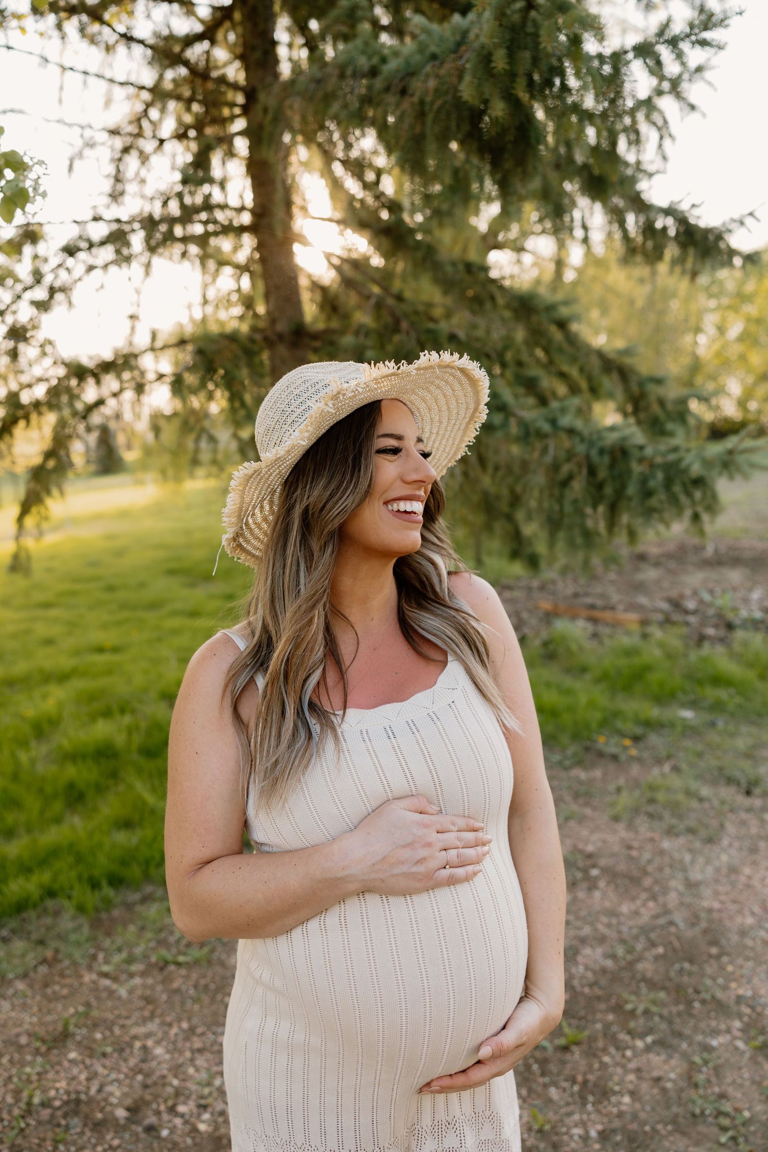 A pregnant woman in a white ribbed dress and straw sun hat stands in front of evergreen trees at sunset.