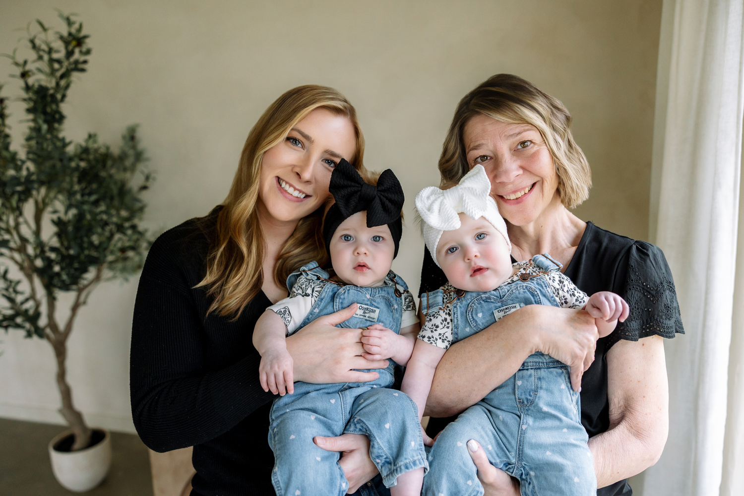 Two women smiling and holding twin baby girls in denim outfits with bow headbands indoors.