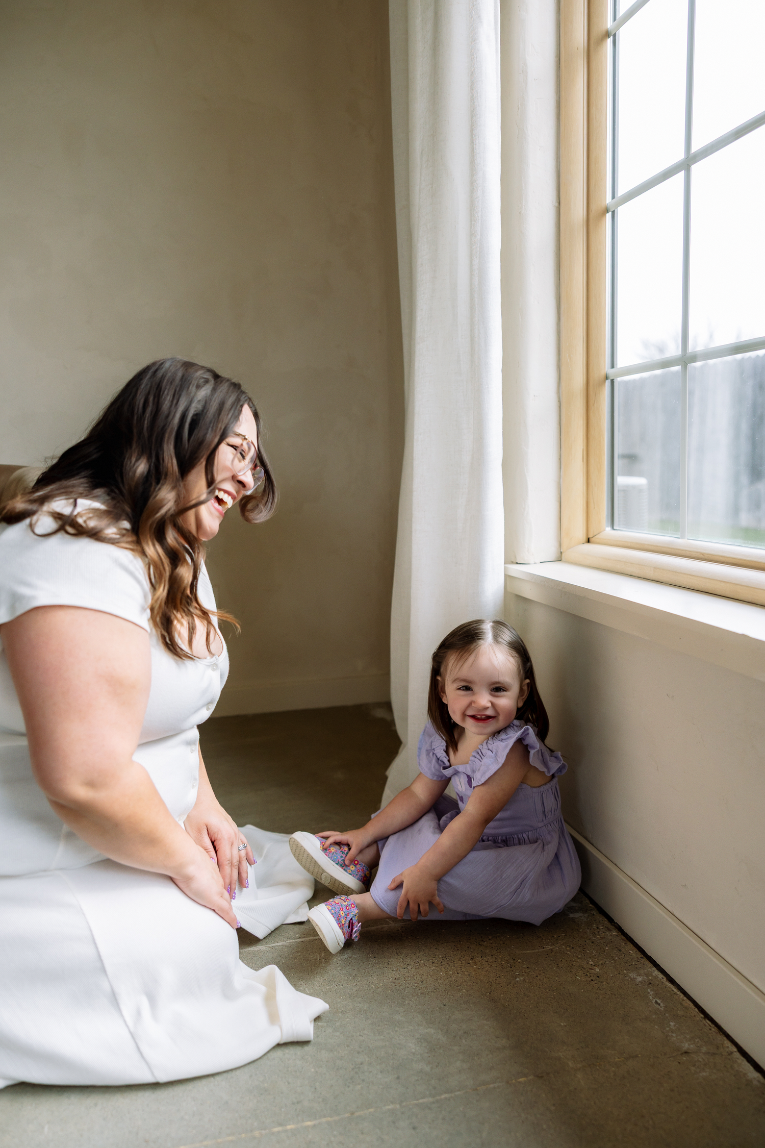 A woman in white dress helps a smiling toddler girl in purple outfit with shoes near a bright window.