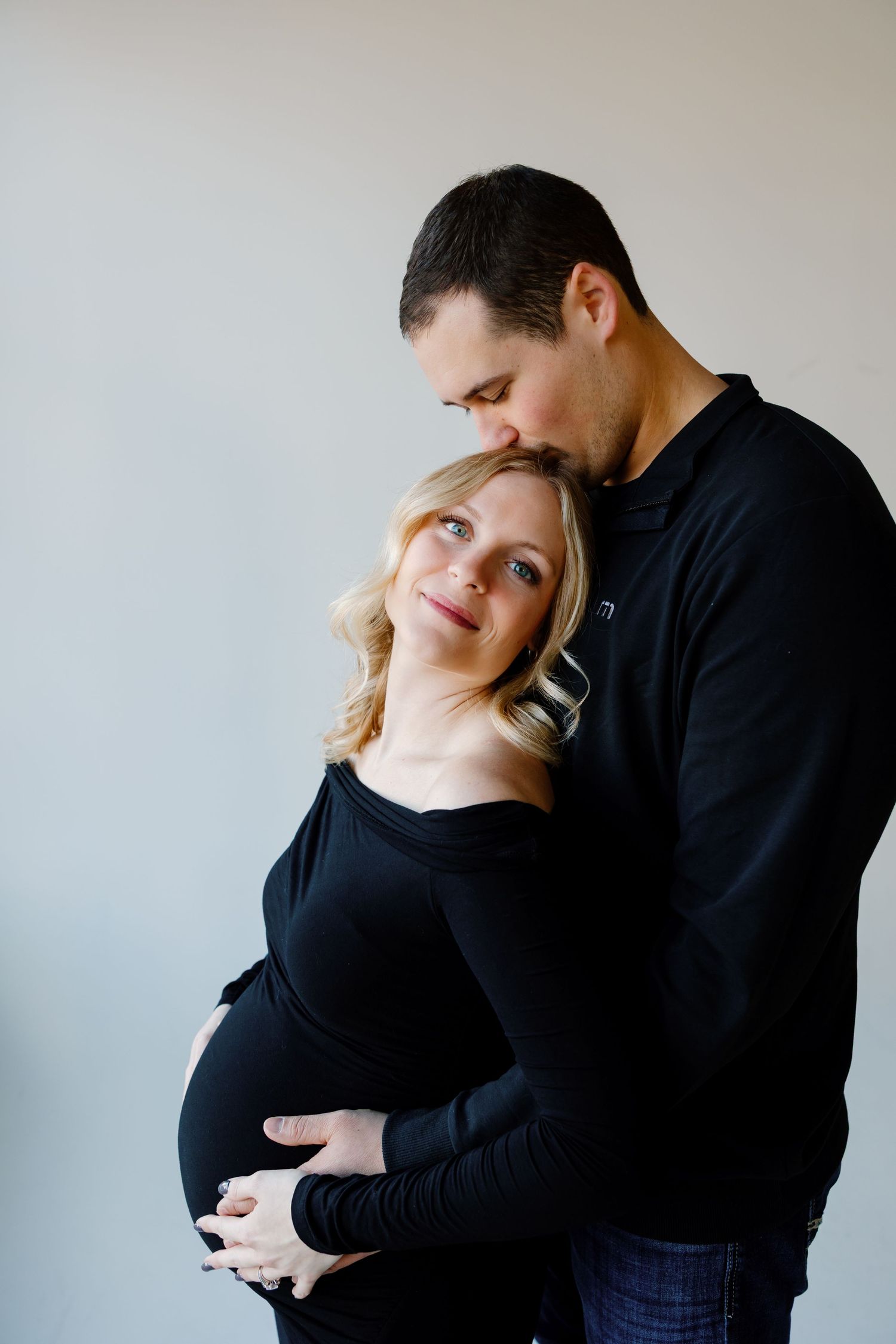 A couple in black clothing embracing during a maternity photoshoot against a light backdrop. Riley Ro Photo Sioux Falls, SD