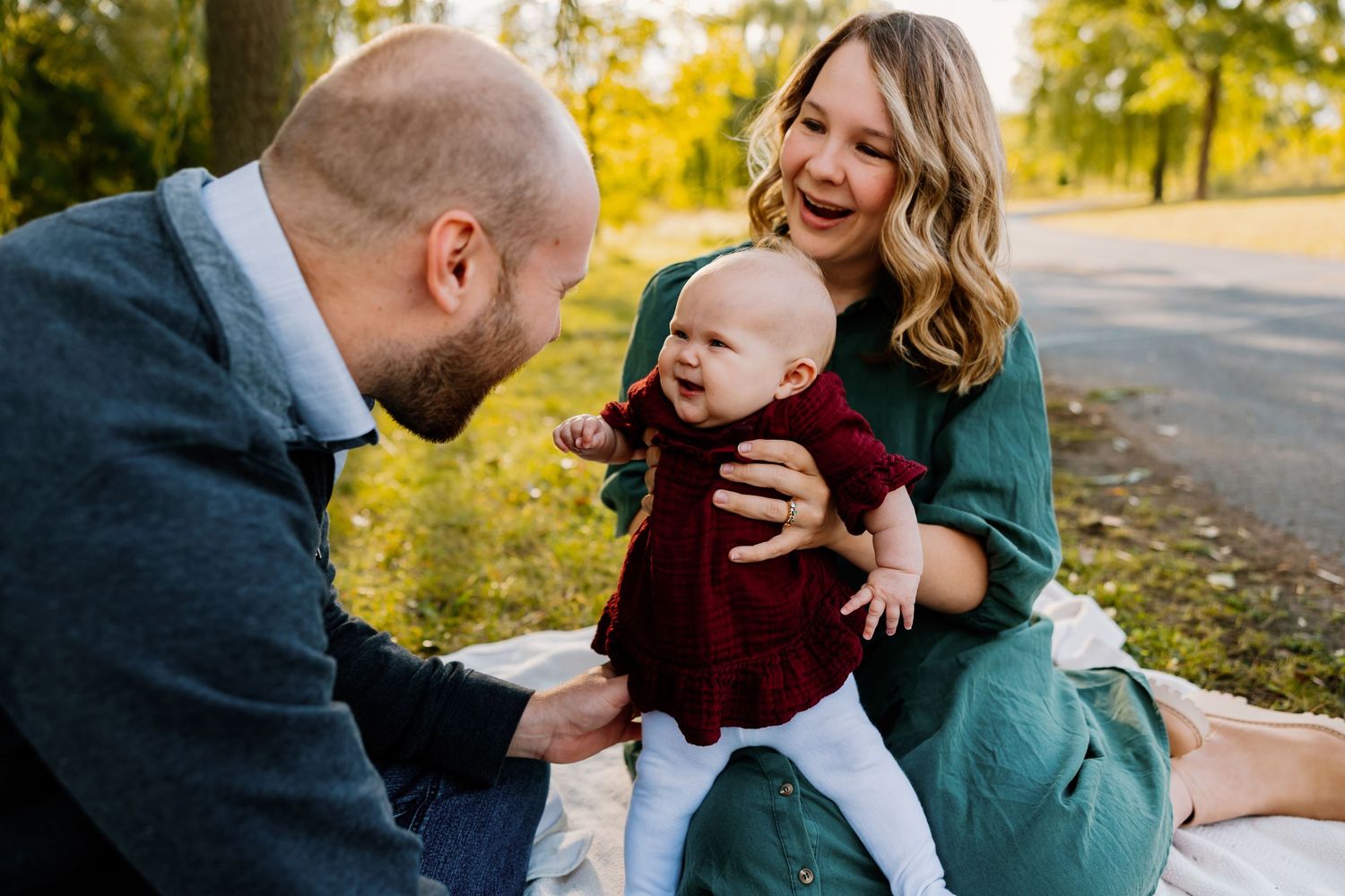 A happy family with a bald dad, smiling mom in green dress, and laughing baby in red outfit sitting outdoors in autumn park.