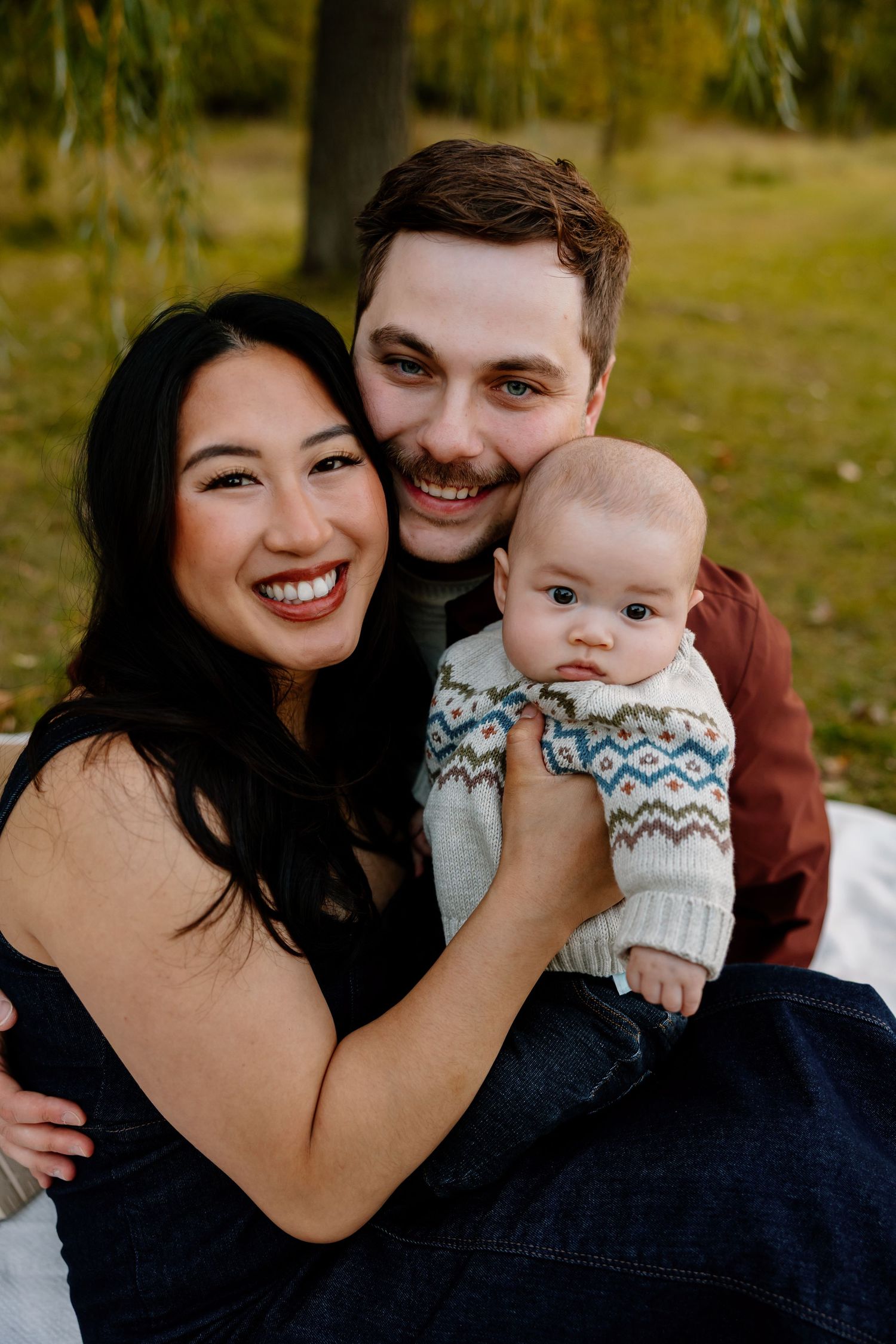 Smiling family with mother, father, and baby in a fair isle sweater posing outdoors in an autumn park setting.