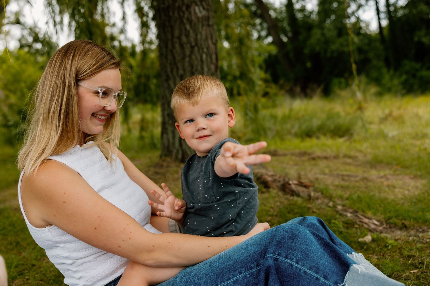 Two people sit together on grass in a wooded outdoor setting on a sunny day.