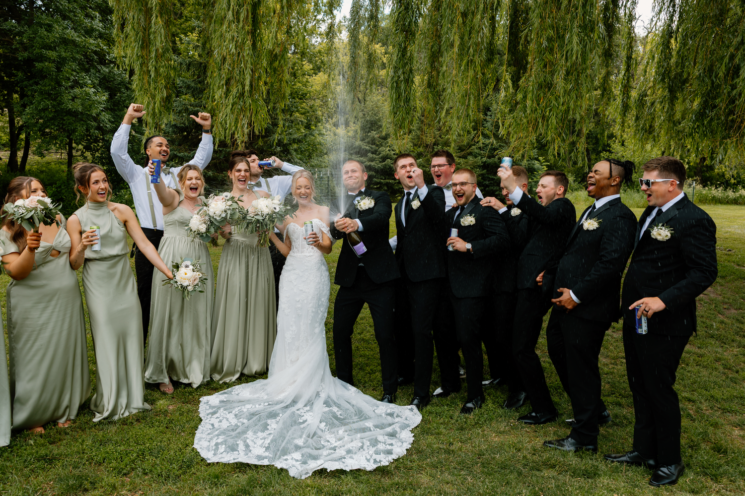 Bride and groom celebrate with full wedding party under willow trees as champagne is sprayed.