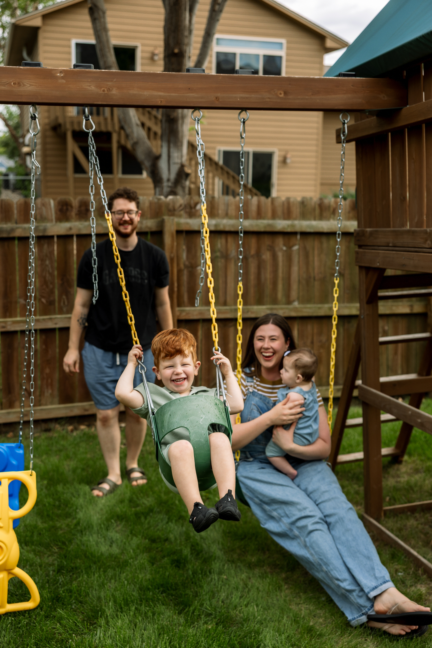 Family enjoying backyard playtime with redhead boy swinging on colorful chain swing set while mom holds baby.
