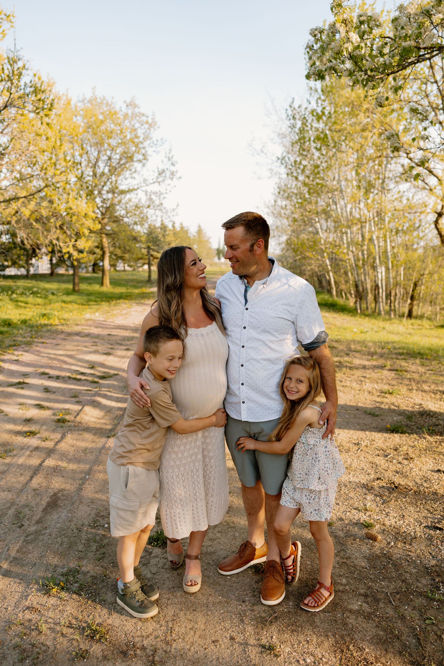 Family dressed in casual attire stands together on dirt path surrounded by golden fall trees in sunlit outdoor setting.