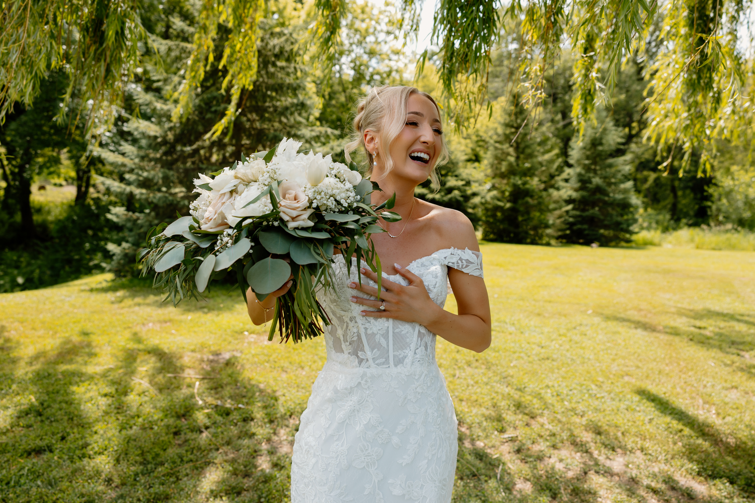 Joyful bride in off-shoulder lace gown holding white floral bouquet, laughing outdoors under weeping willow trees.