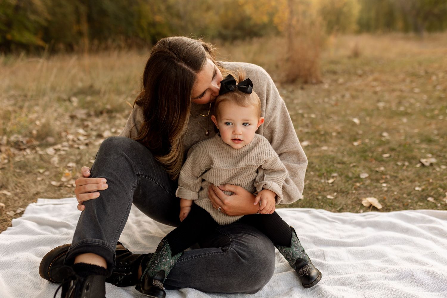 A mother sits on a white blanket outdoors, lovingly holding her baby girl in a cozy autumn field setting.