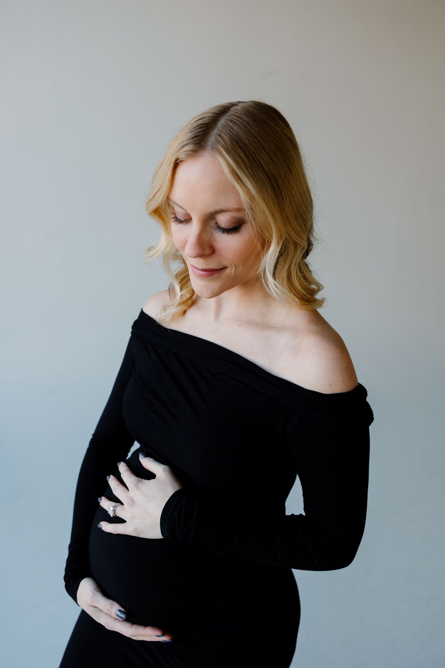 A pregnant woman in a black off-shoulder dress poses elegantly against a neutral background. Riley Ro Photo Sioux Falls, SD