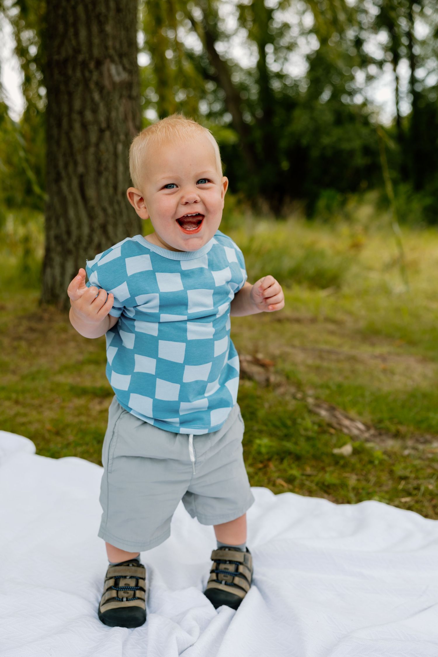 A toddler wearing a blue checkered shirt and gray shorts laughs joyfully while standing on a white blanket outdoors.