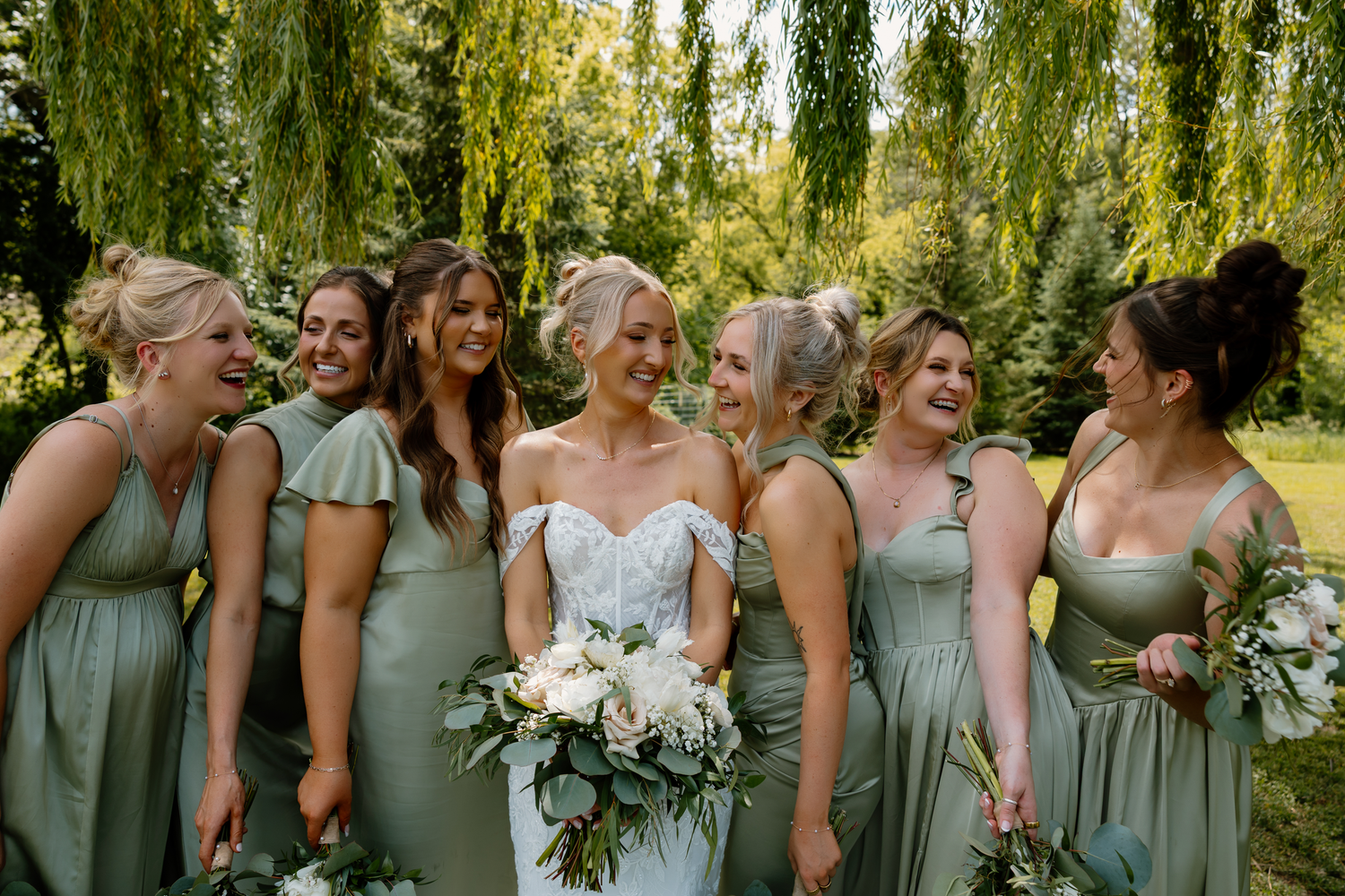 Bride in white gown with bridesmaids in sage green dresses holding bouquets under willow trees.