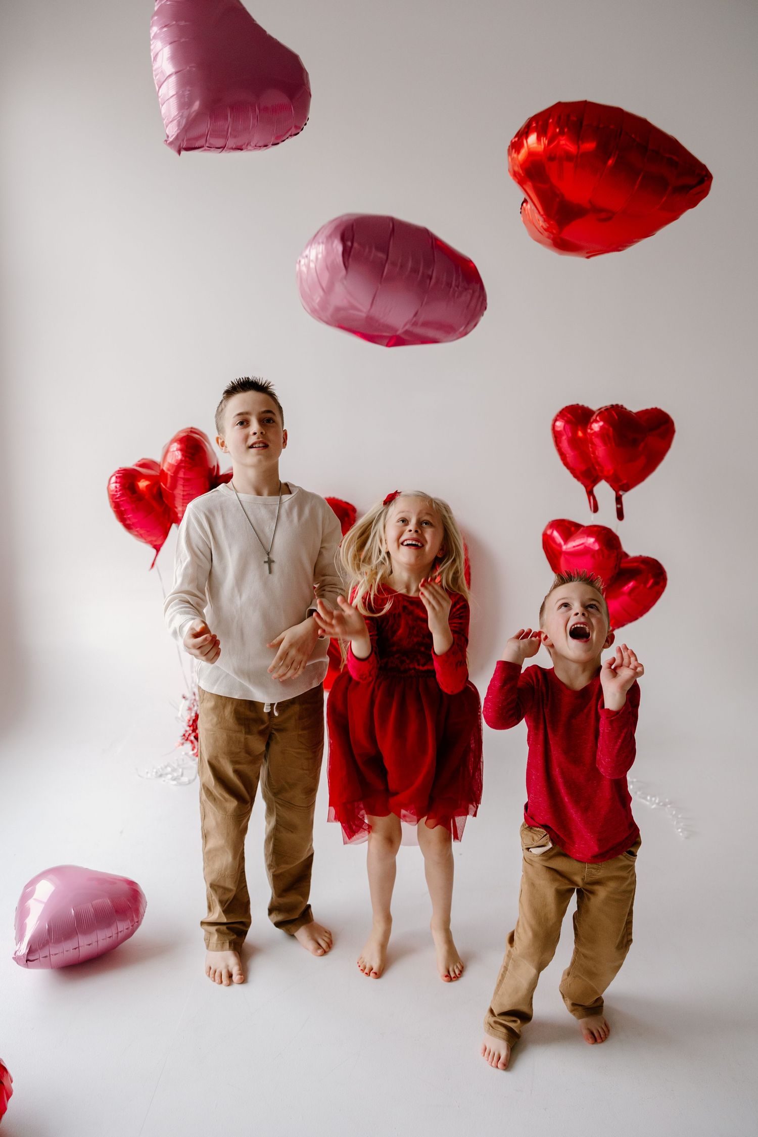 Children playing with pink and red heart-shaped balloons against white wall during Valentine's day photoshoot.
