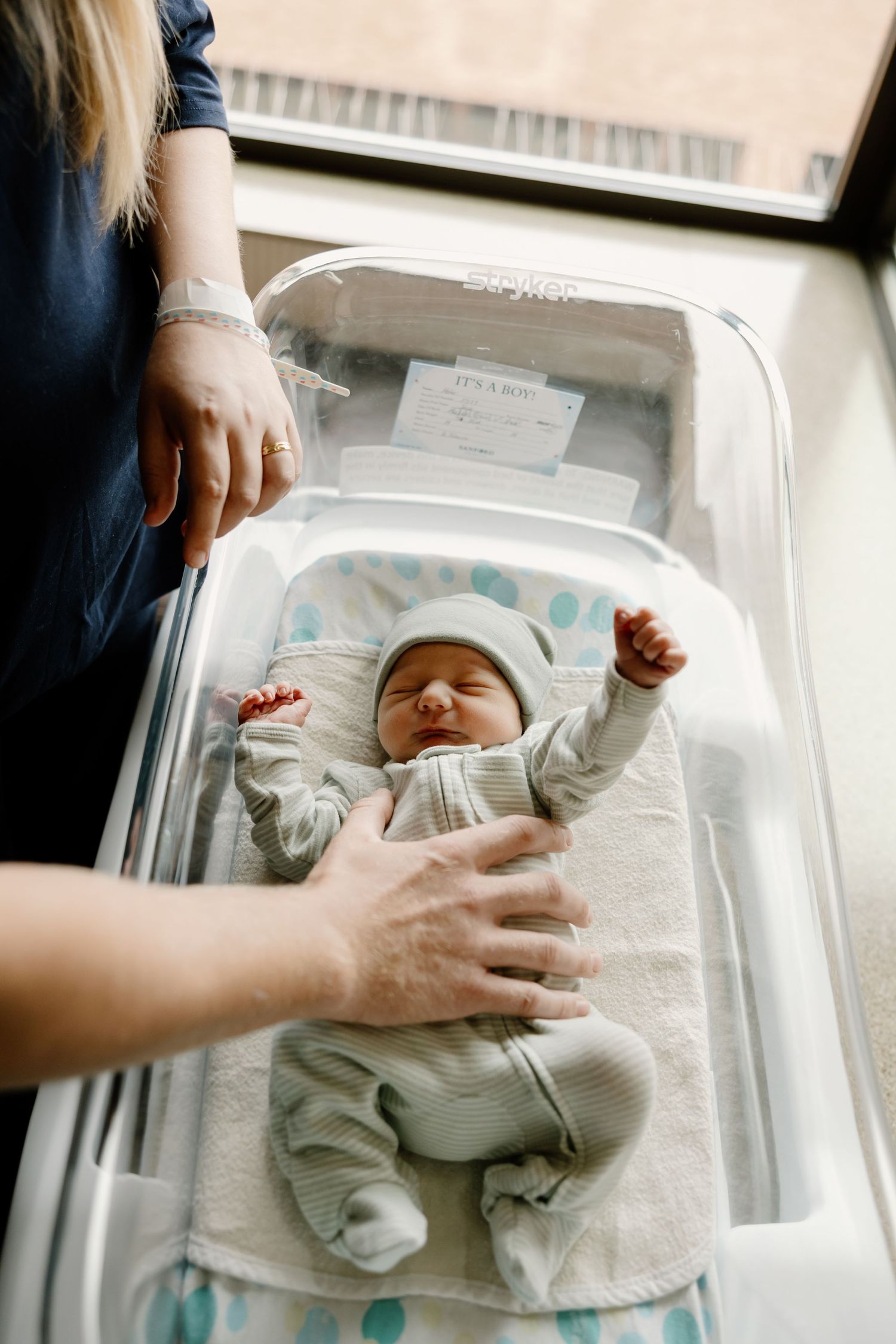 A newborn baby rests in a hospital bassinet as gentle hands provide care.