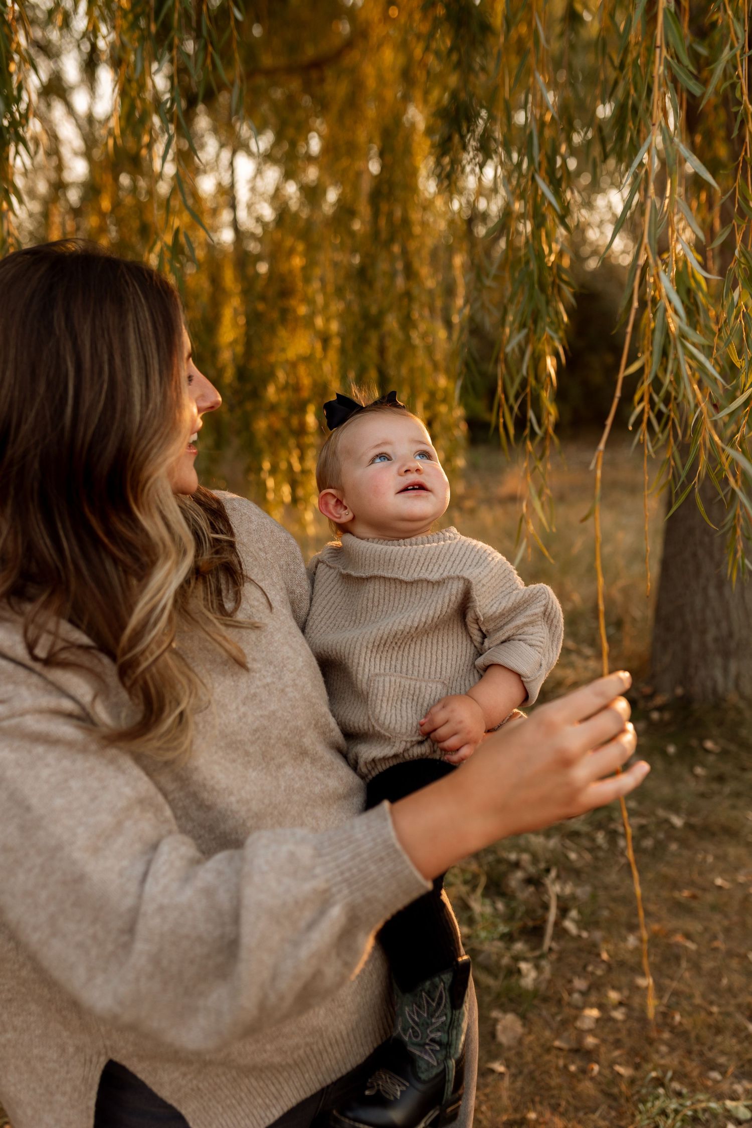 Mother holding smiling baby under willow tree in golden autumn light, both wearing cozy beige sweaters.