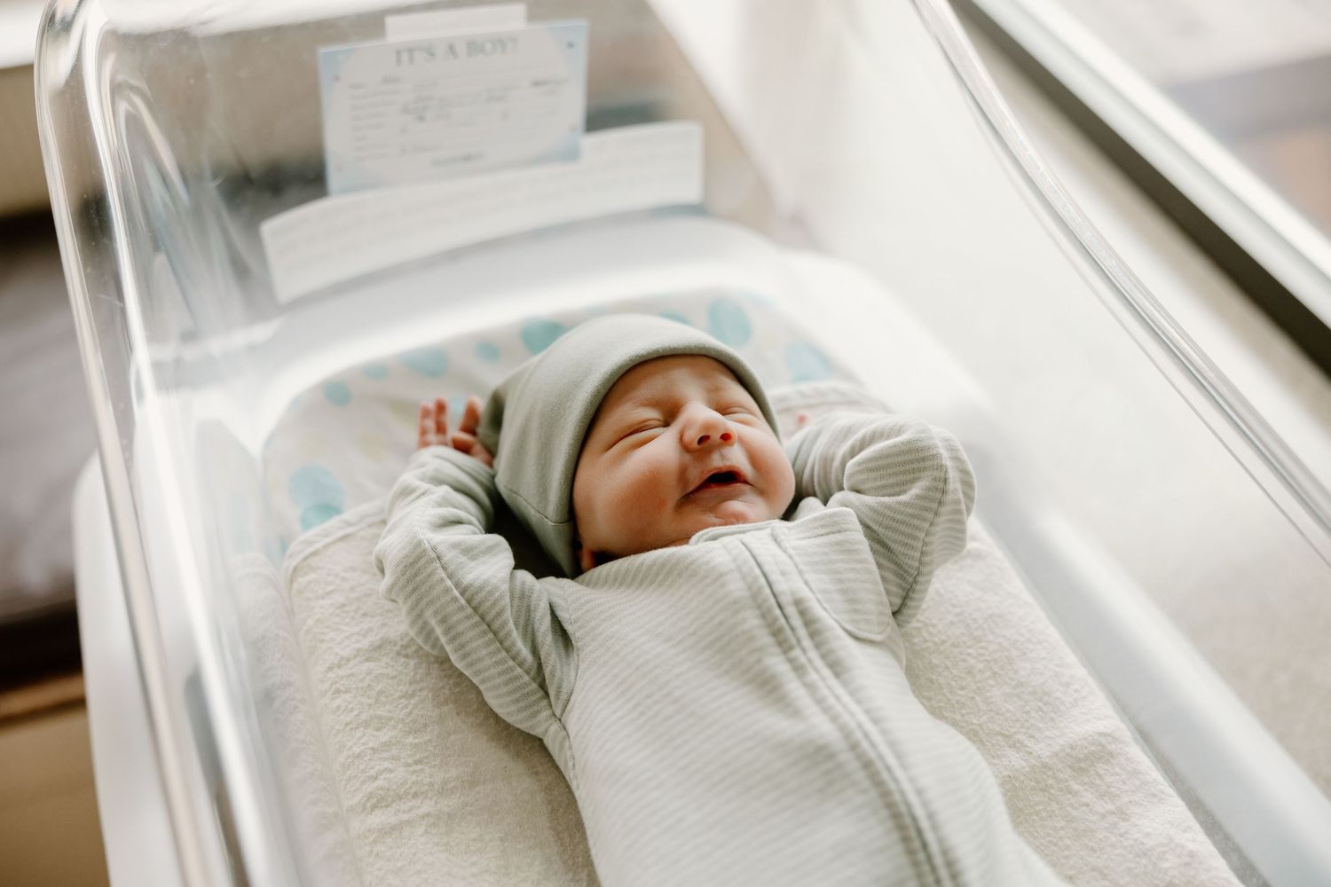 Newborn baby in white zippered sleeper rests in a hospital bassinet with polka dot bedding.