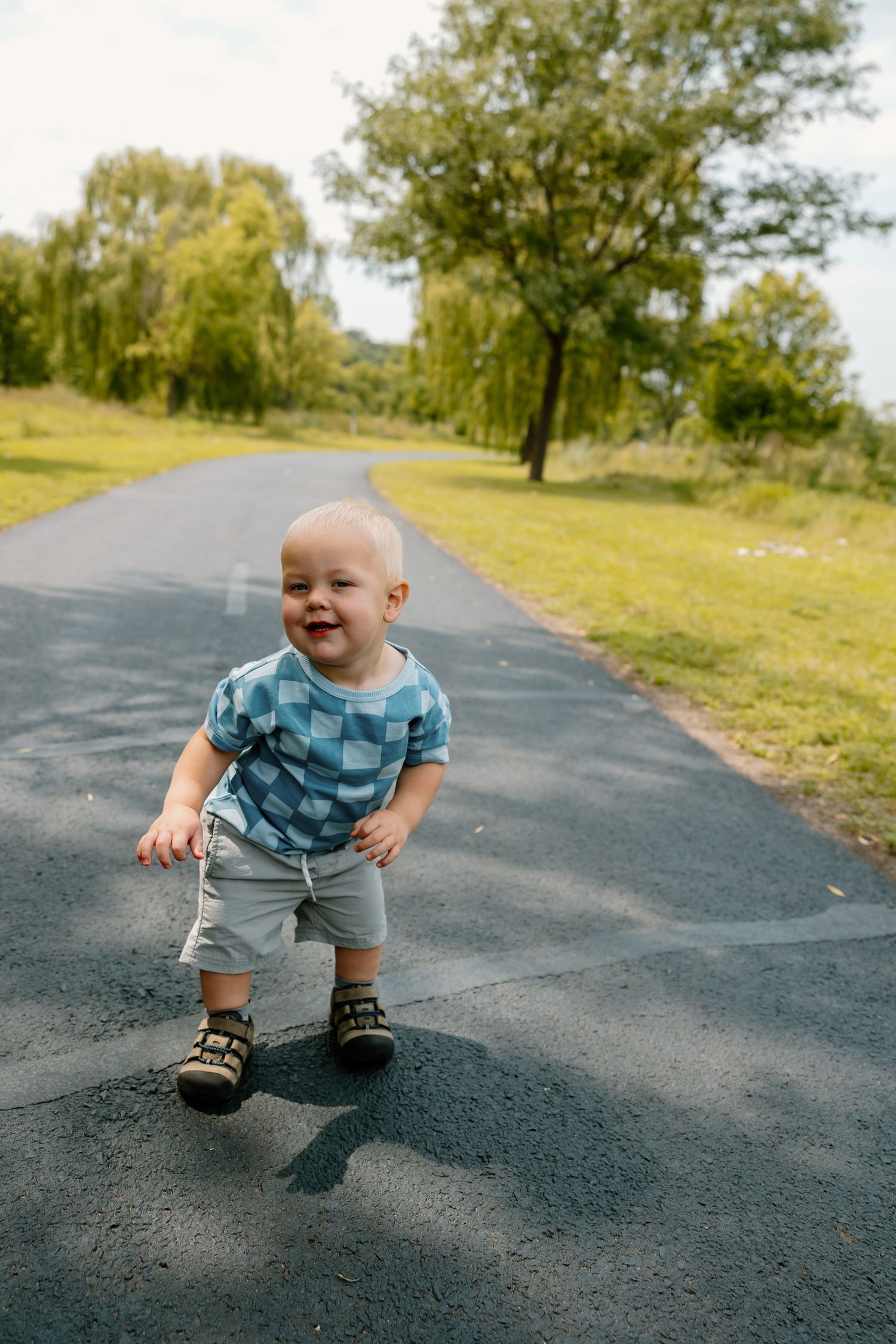 Toddler in a blue checkered shirt walking on a paved path surrounded by green trees.