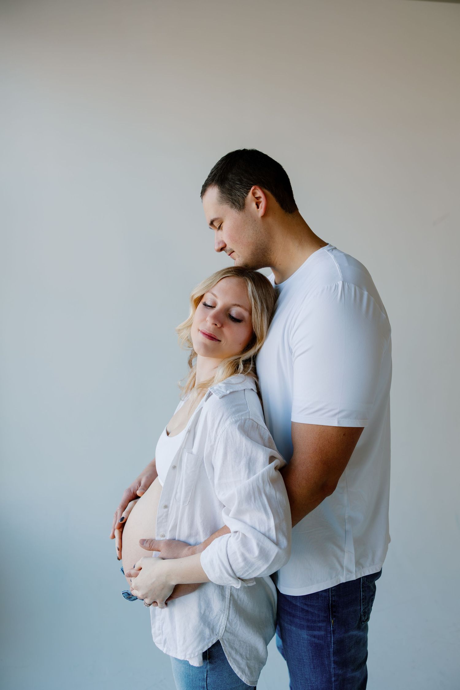 An intimate maternity portrait against a light gray backdrop shows a tender embrace. Riley Ro Photo Sioux Falls, SD