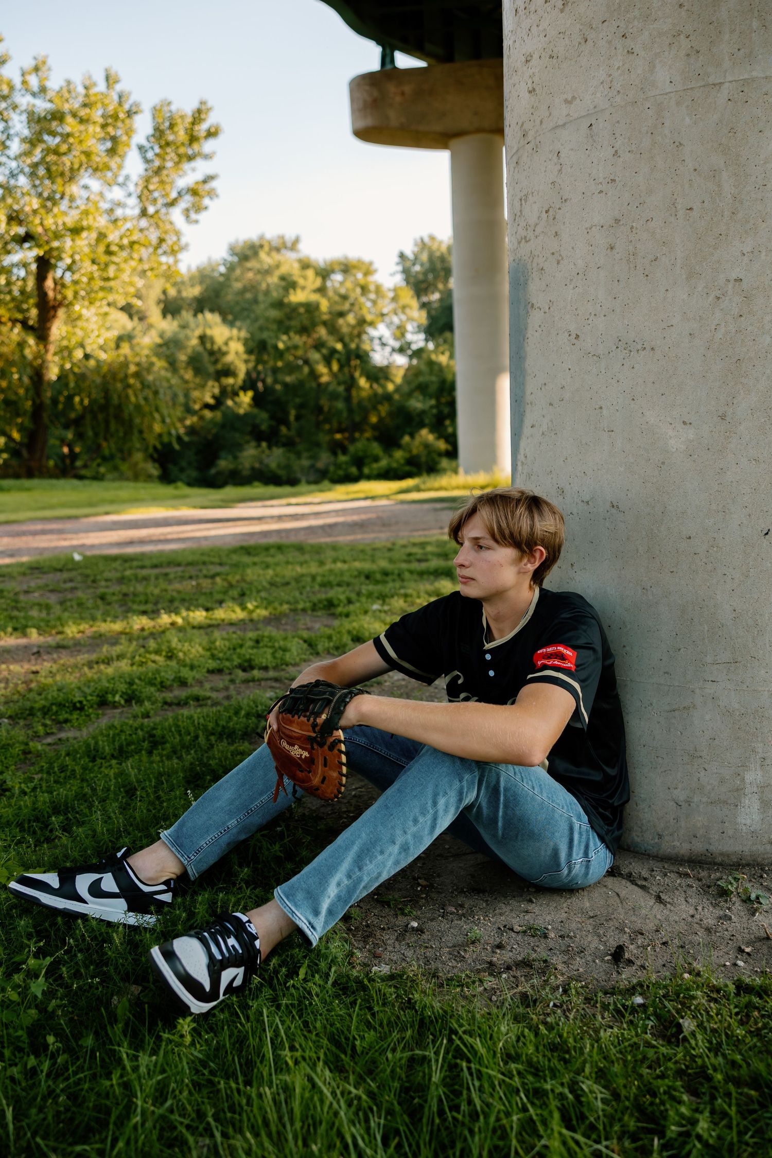 Young athlete in black jersey and jeans relaxes against concrete pillars during outdoor photoshoot.
