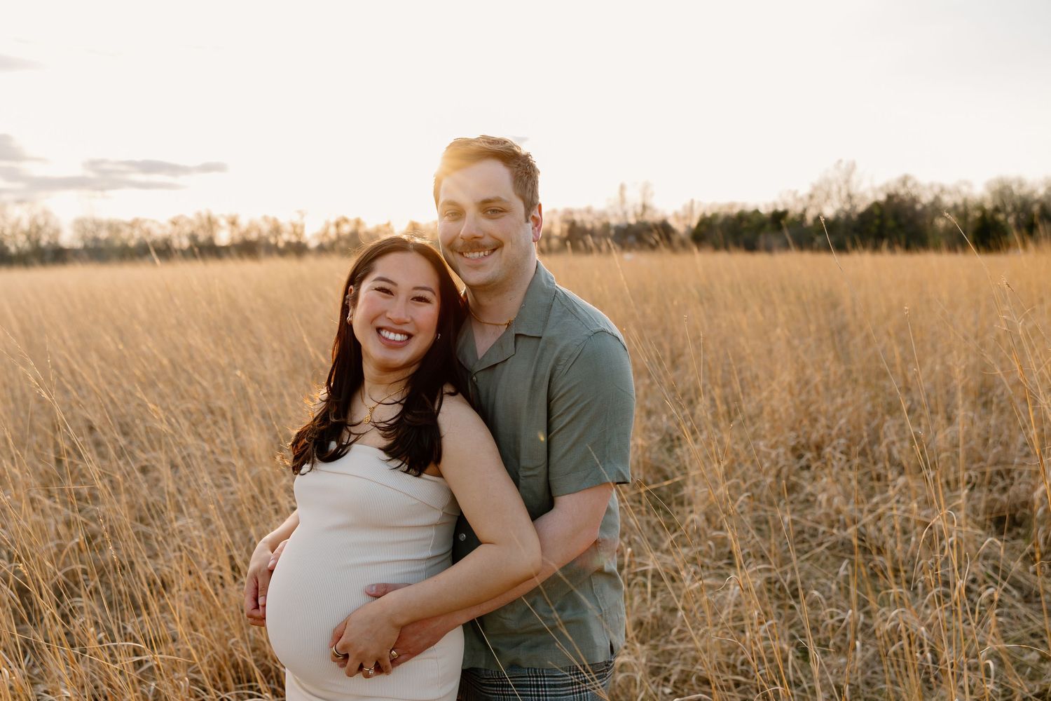 Joyful expectant couple embracing in sunlit field during sunset maternity photo session.