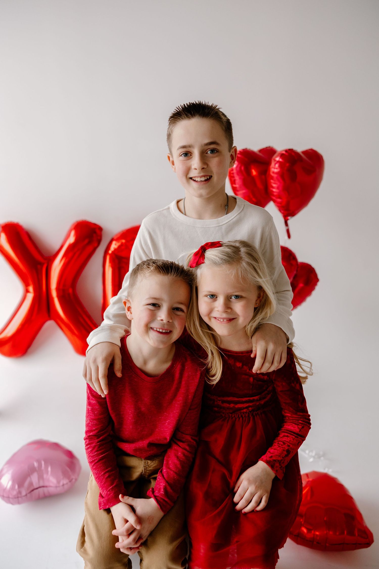 Three young children pose together against a white wall decorated with red heart-shaped balloons and an X decoration.