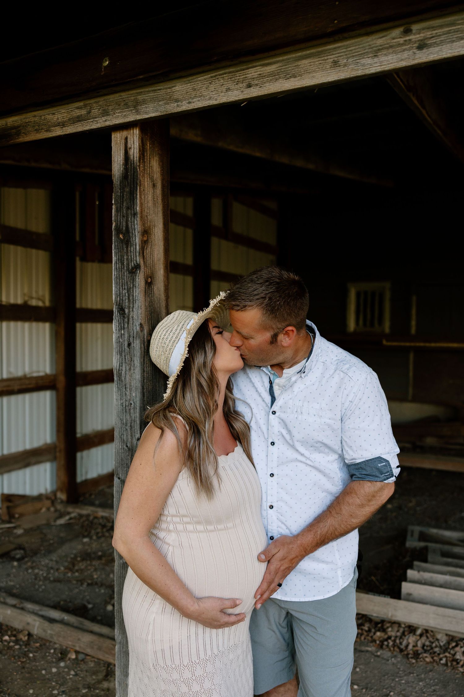 A couple shares a tender moment in a rustic barn setting with the woman wearing a flowing white dress and sun hat.