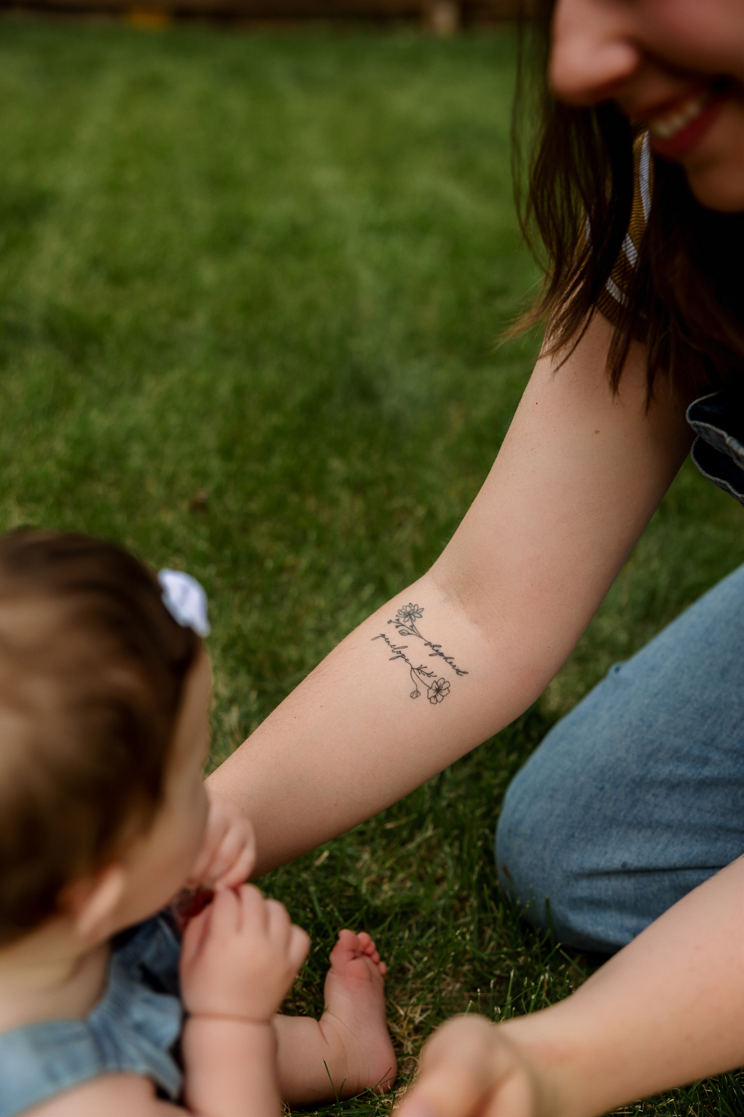 Close-up of a mother's forearm tattoo as her baby reaches toward her while sitting on green grass.