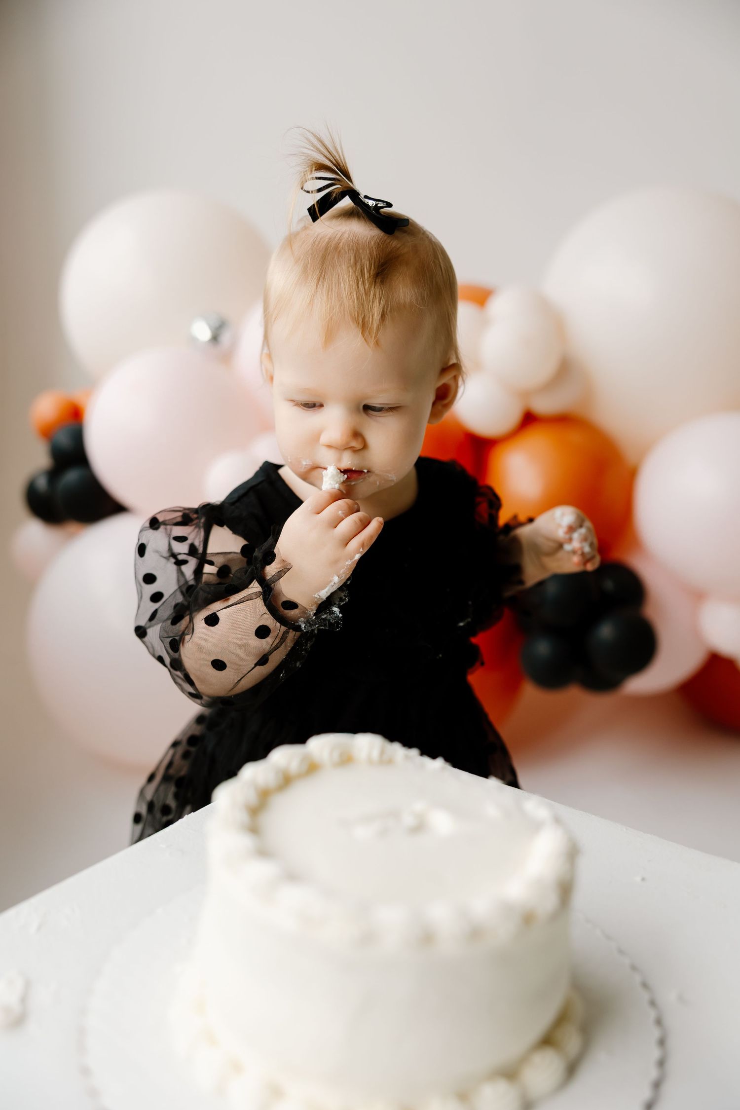 A small child in black polka dot dress tastes frosting near a white cake with balloon decorations in background.