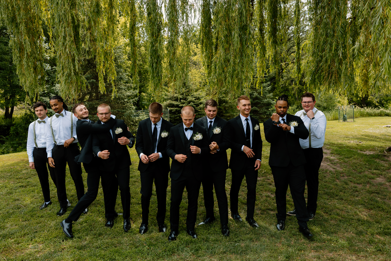 Groomsmen in black suits laughing and celebrating together under lush weeping willow trees at a wedding.