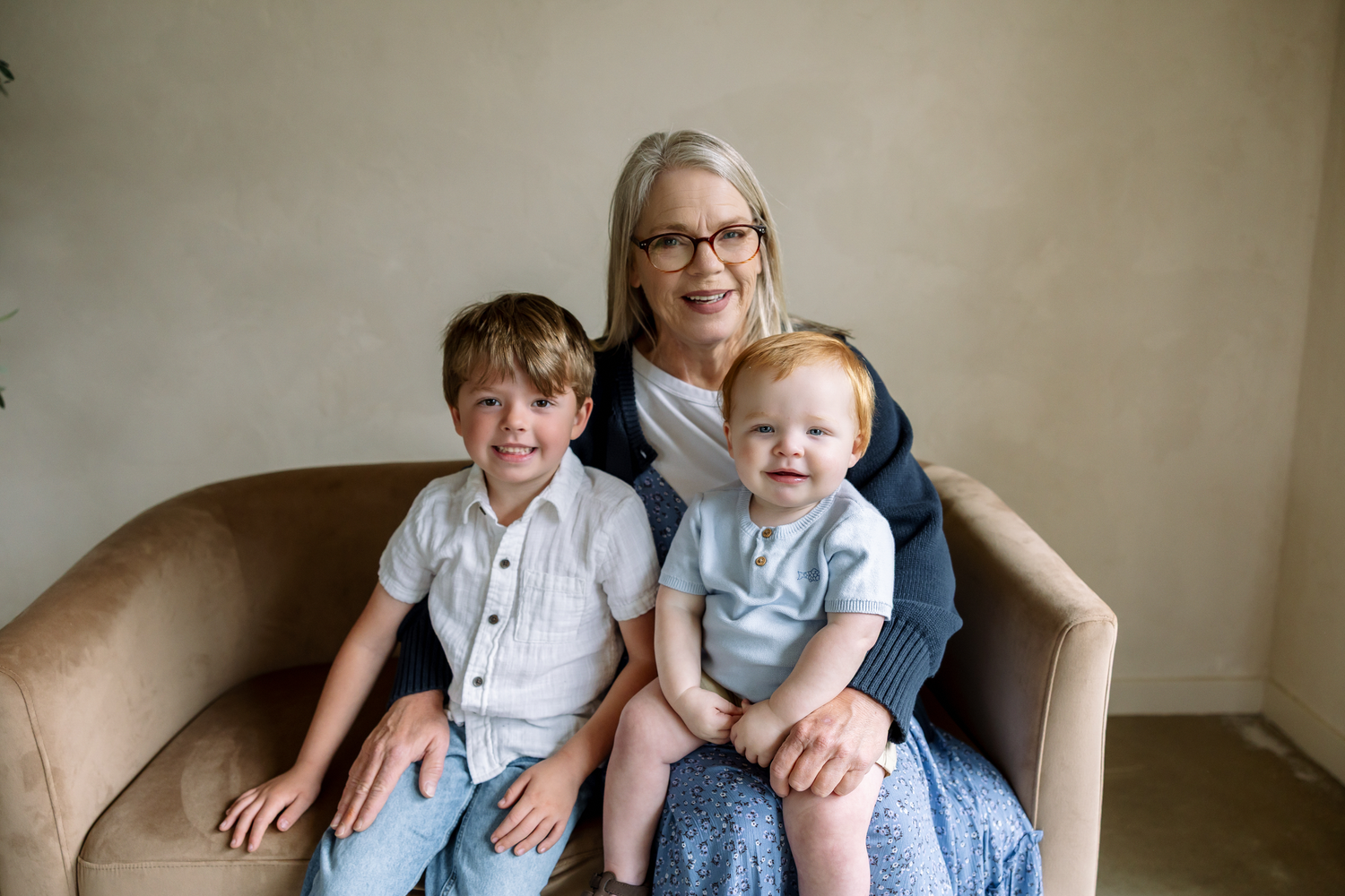 Grandmother smiling with two young boys sitting on a beige armchair in a bright, neutral-toned room.