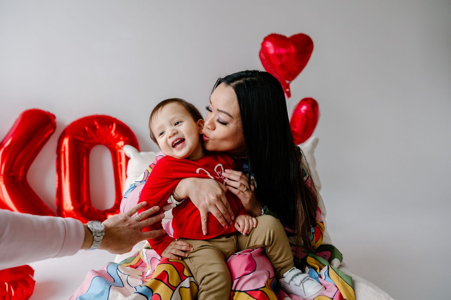 A sweet moment captured against grey wall with colorful blankets and red heart-shaped balloons.
