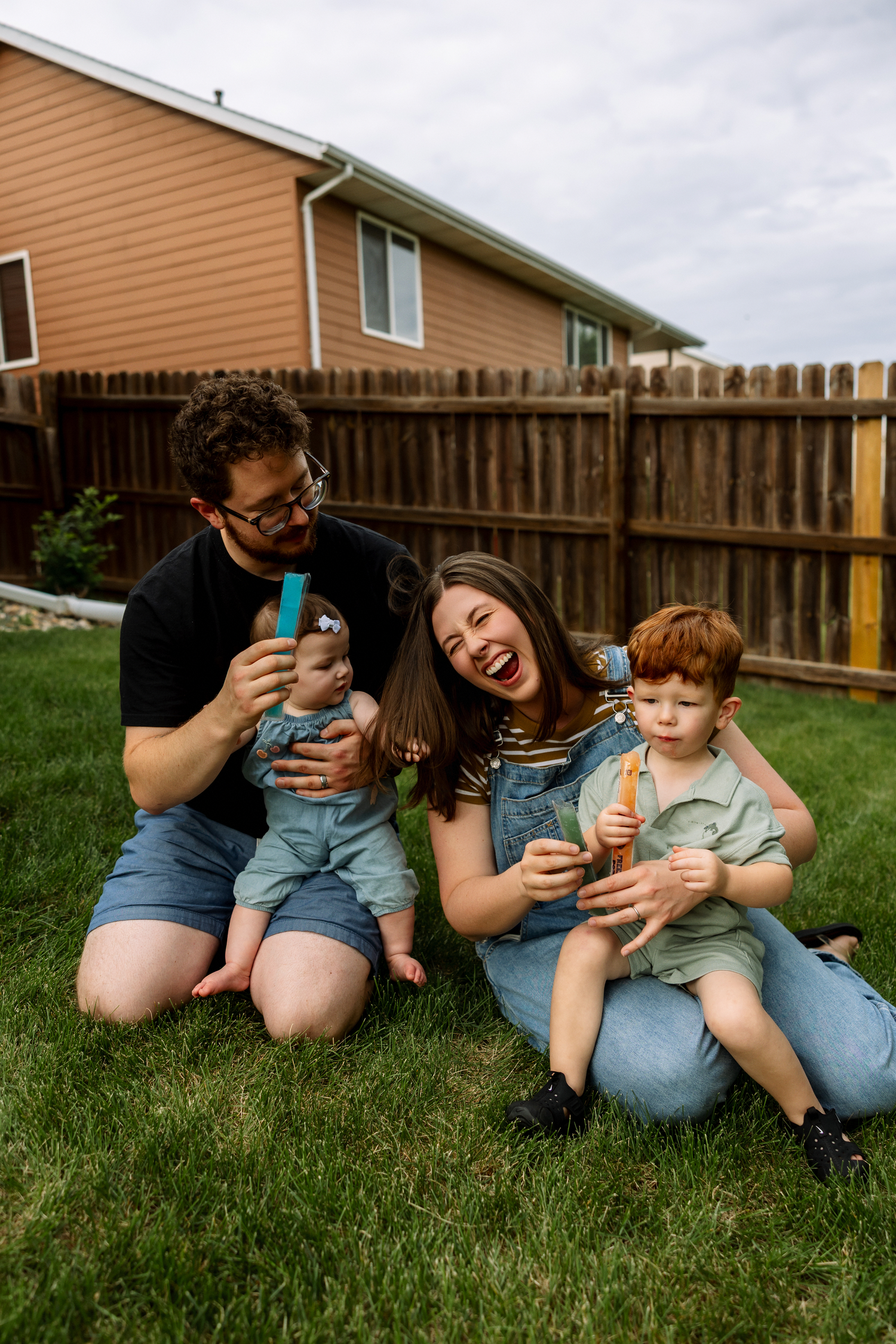 Family of four sitting on backyard grass, parents laughing while blowing bubbles with two young children on a cloudy day.
