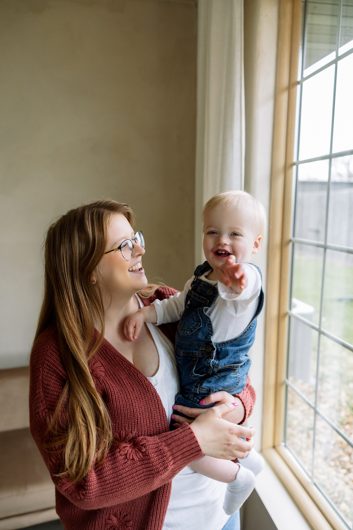 A smiling mother with glasses holds her laughing baby in denim overalls near a sunlit window.