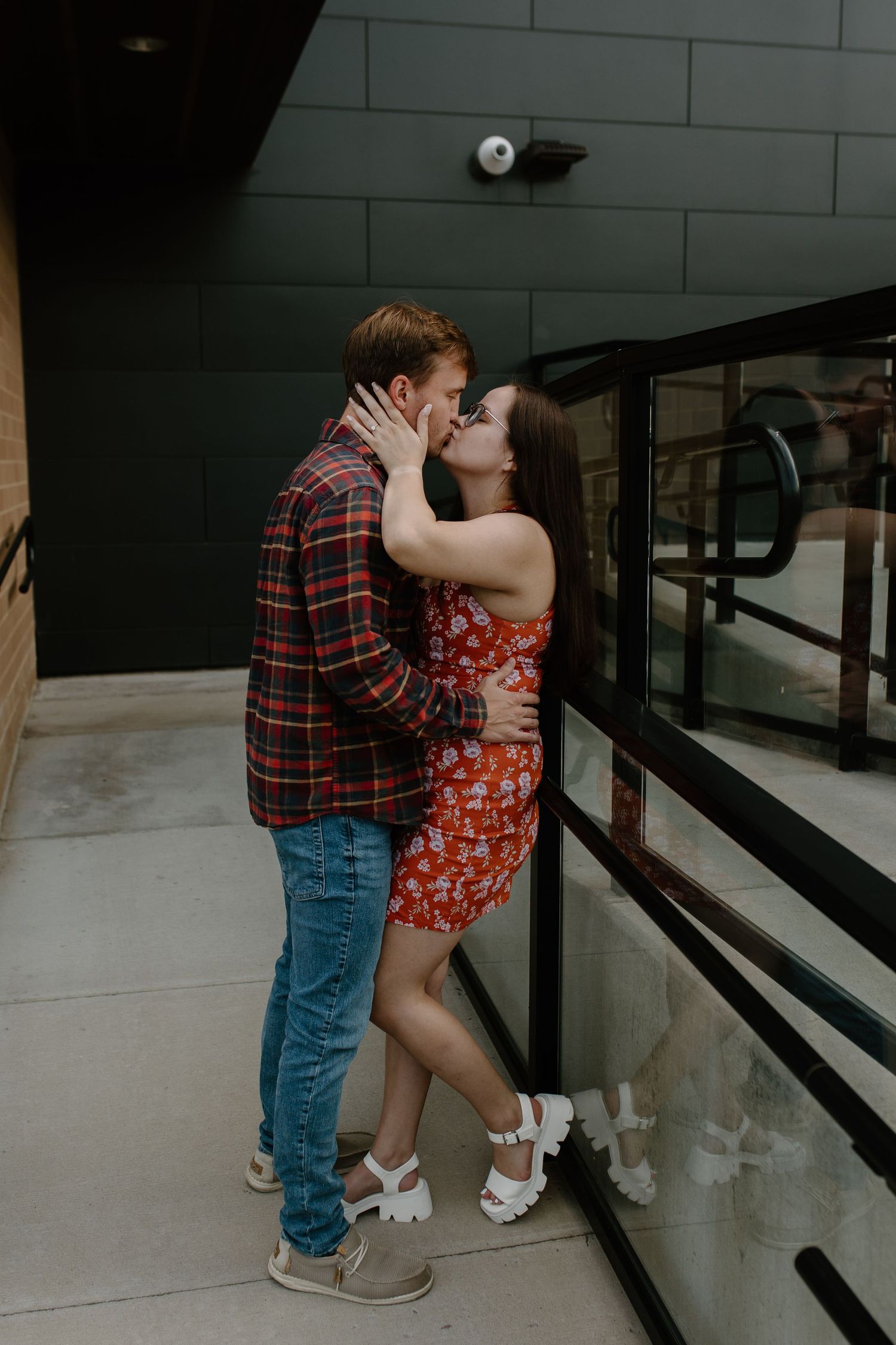 A couple sharing a romantic kiss in an urban setting, with the woman wearing a red floral dress and the man in a plaid shirt.