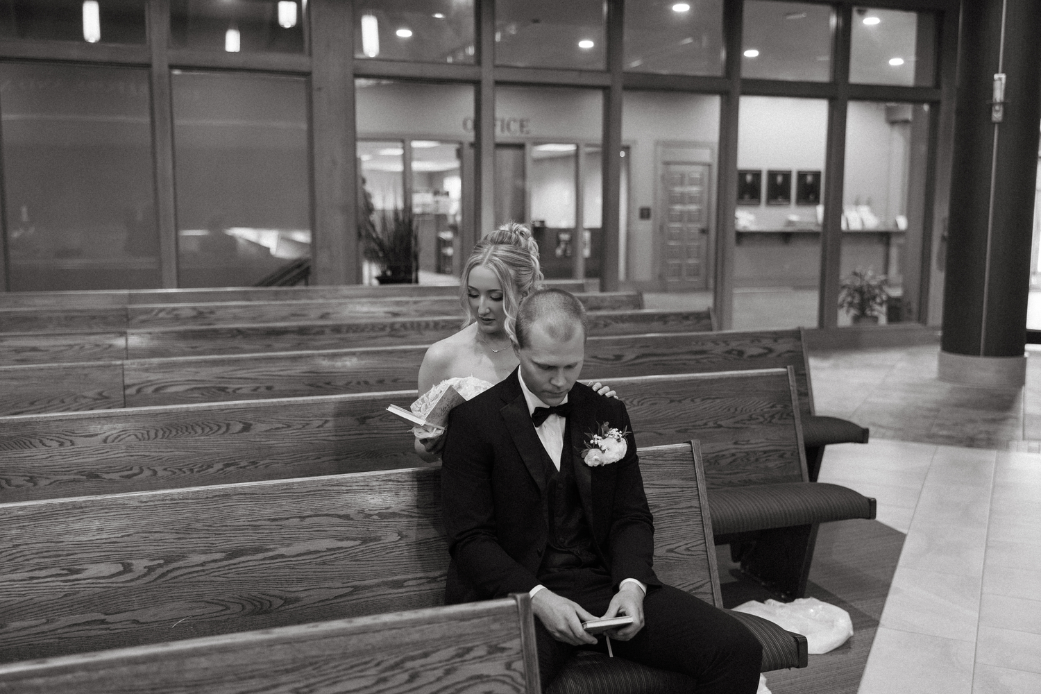 Groom in black tuxedo sits on church pew checking phone while bride stands behind him smiling.