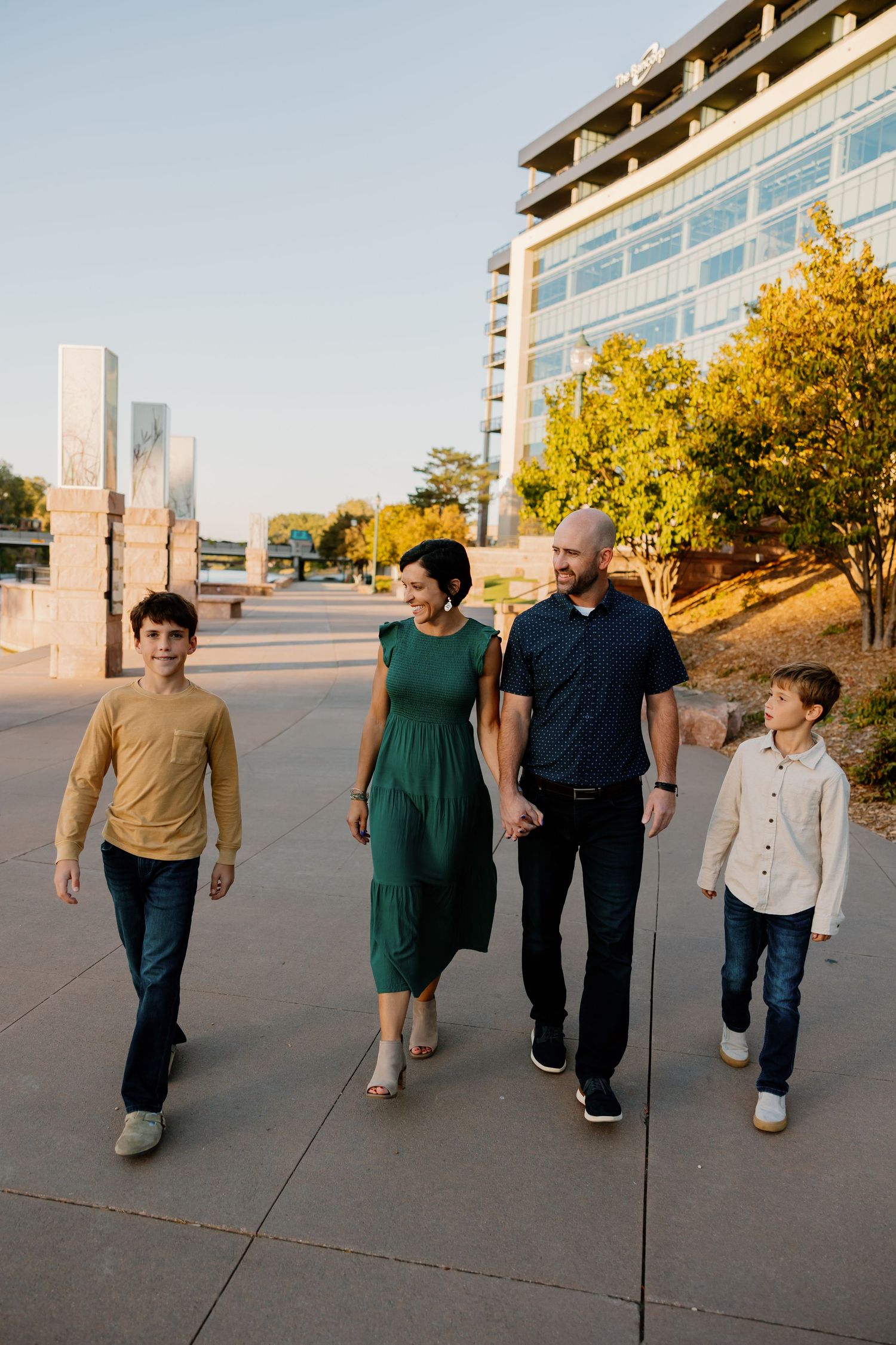 Family walking together on urban sidewalk at sunset with modern building and autumn trees in background. Riley Ro Photo Sioux Falls, SD