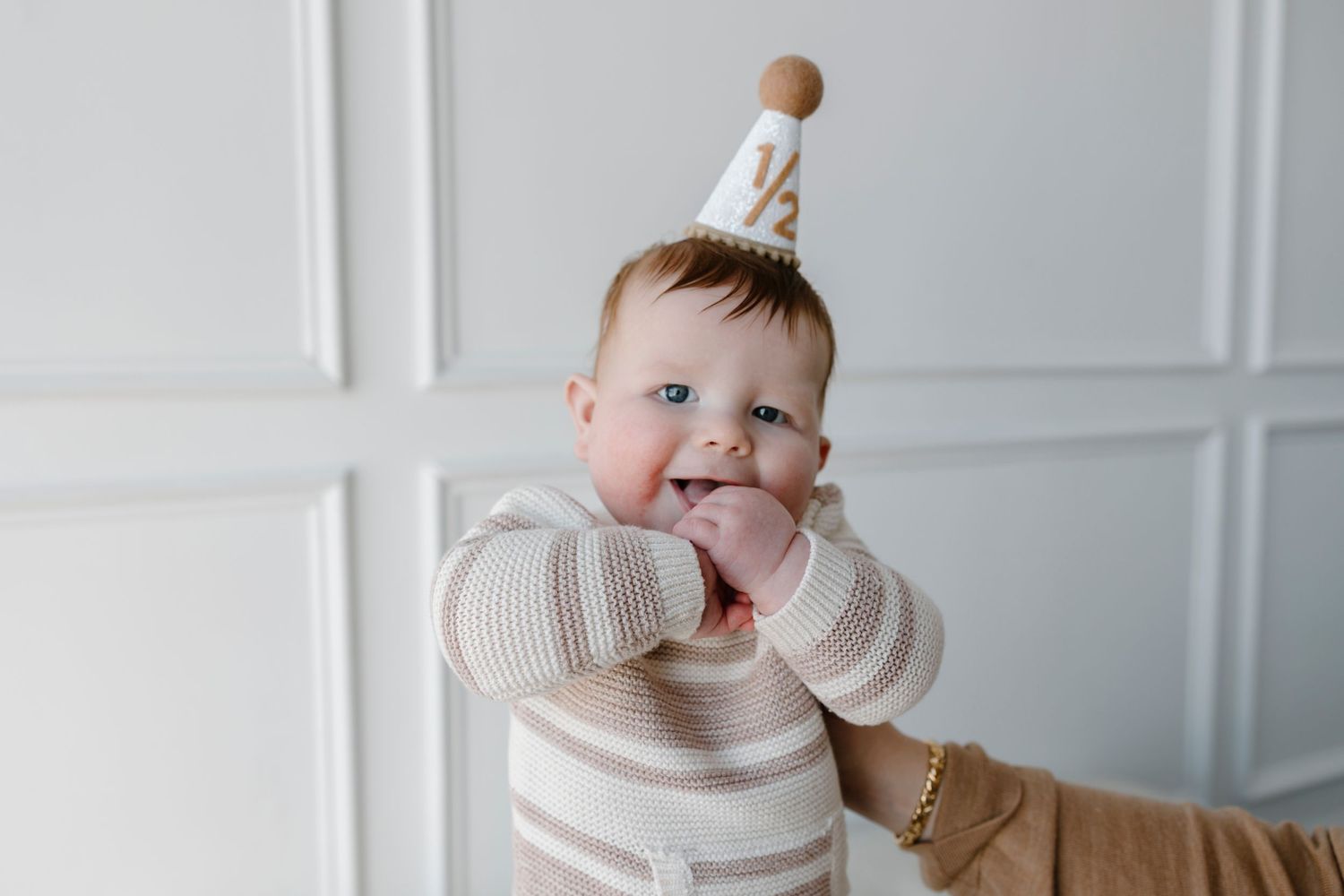 Baby in a white crown sits on a white chair in an elegant room with white paneled walls.