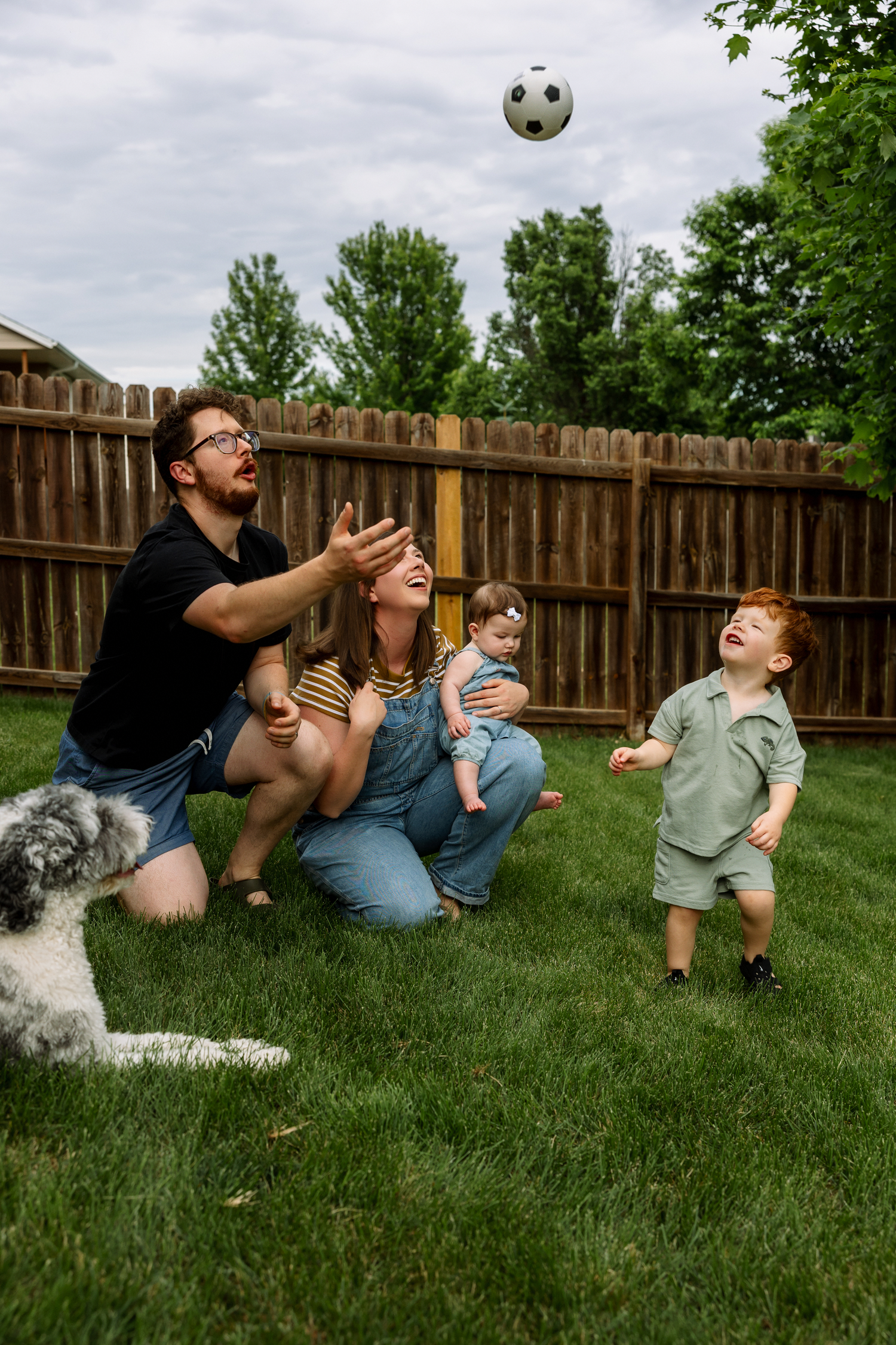 Family playing with a soccer ball in a backyard, with two children, a baby, and a gray fluffy dog.