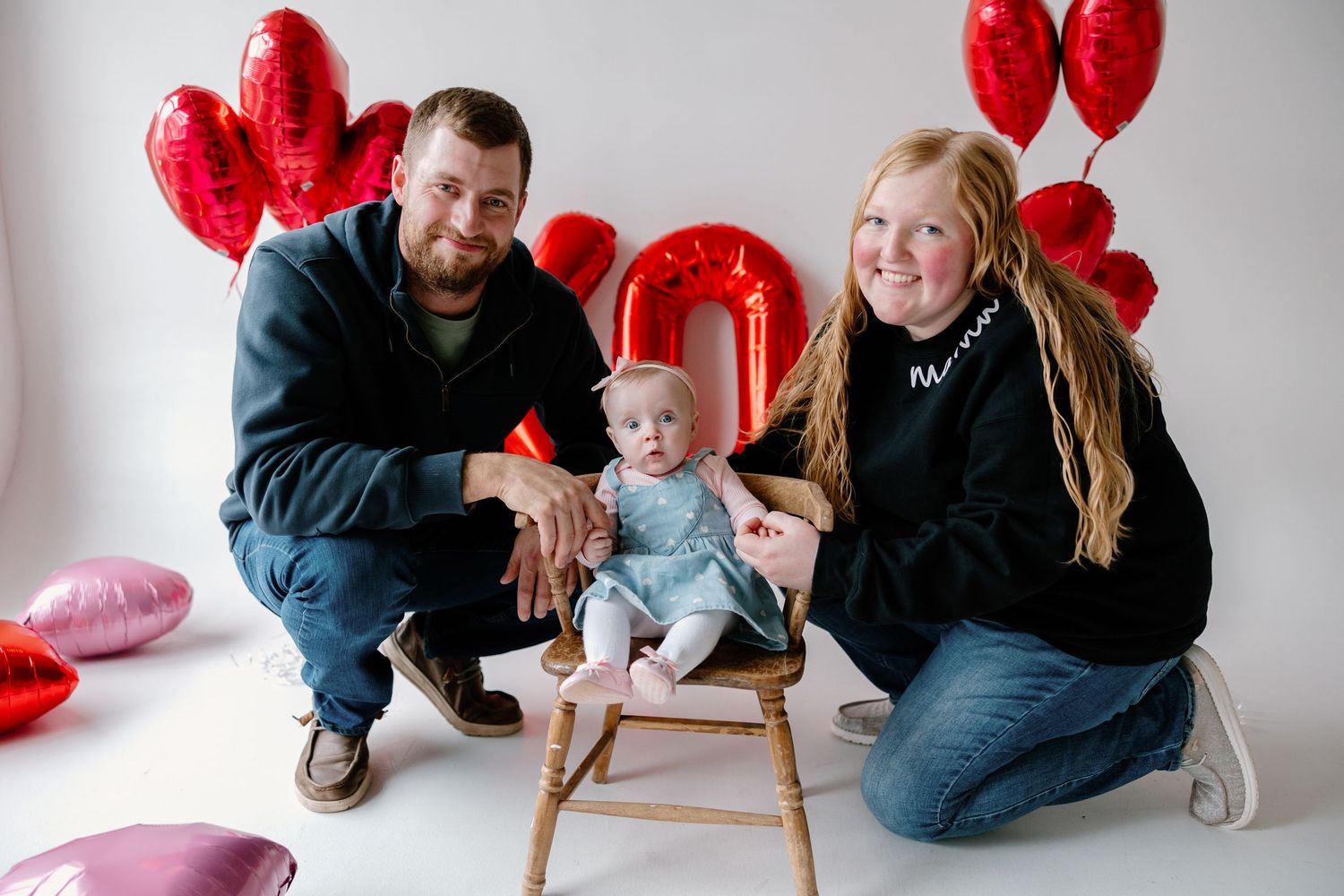 Family portrait with red heart balloons and baby sitting in wooden chair against white background.
