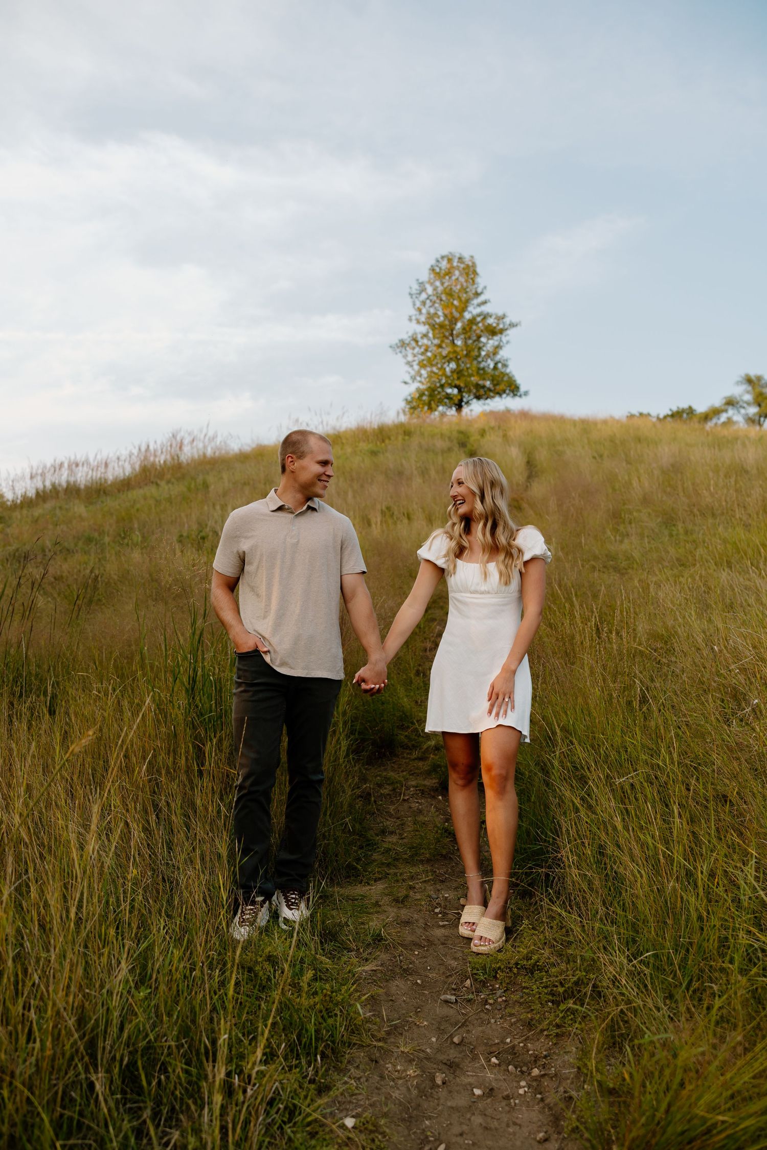 Couple holding hands while walking along a dirt trail through tall grass.