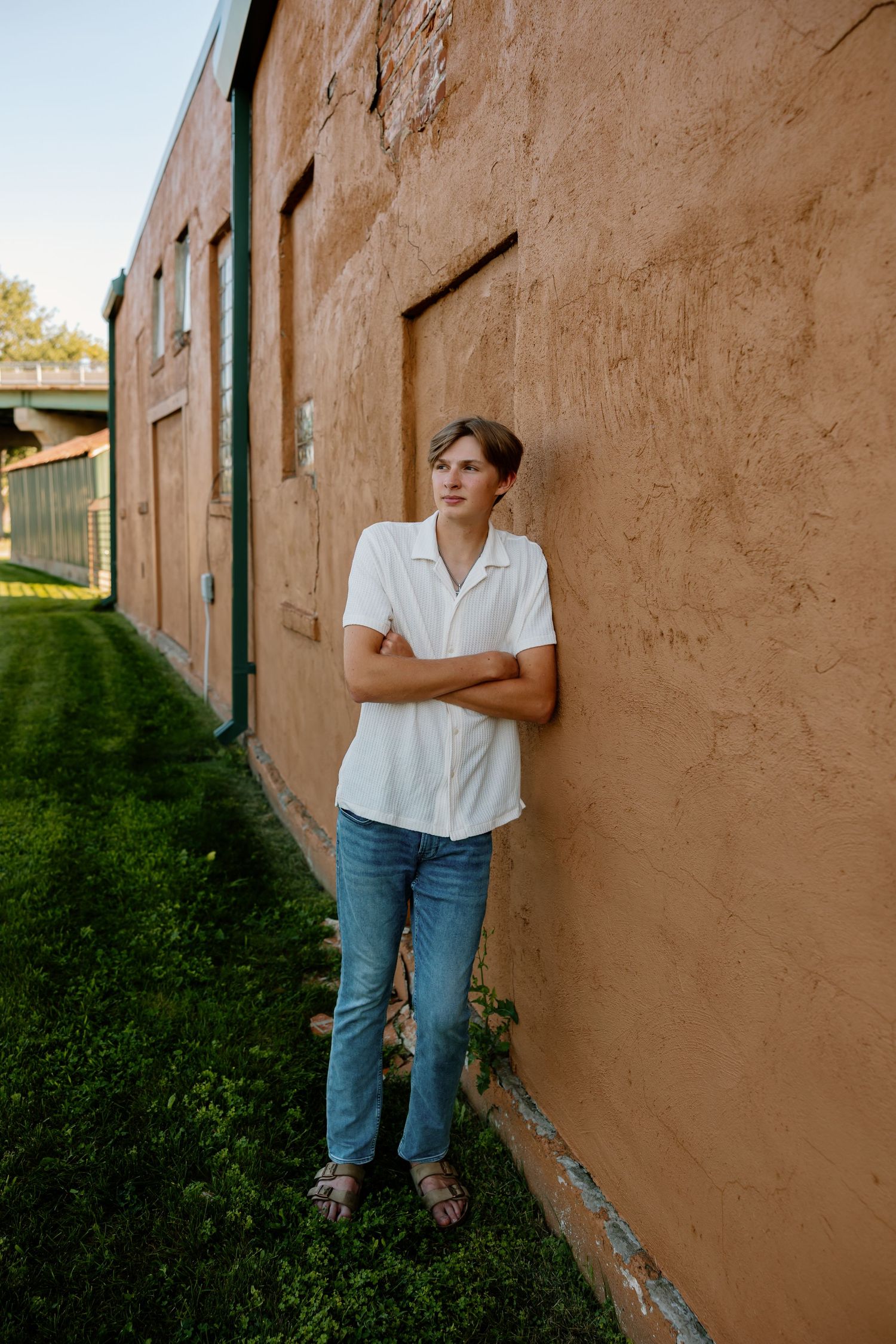 Person in white shirt and jeans standing against an adobe wall with green grass and soft lighting.