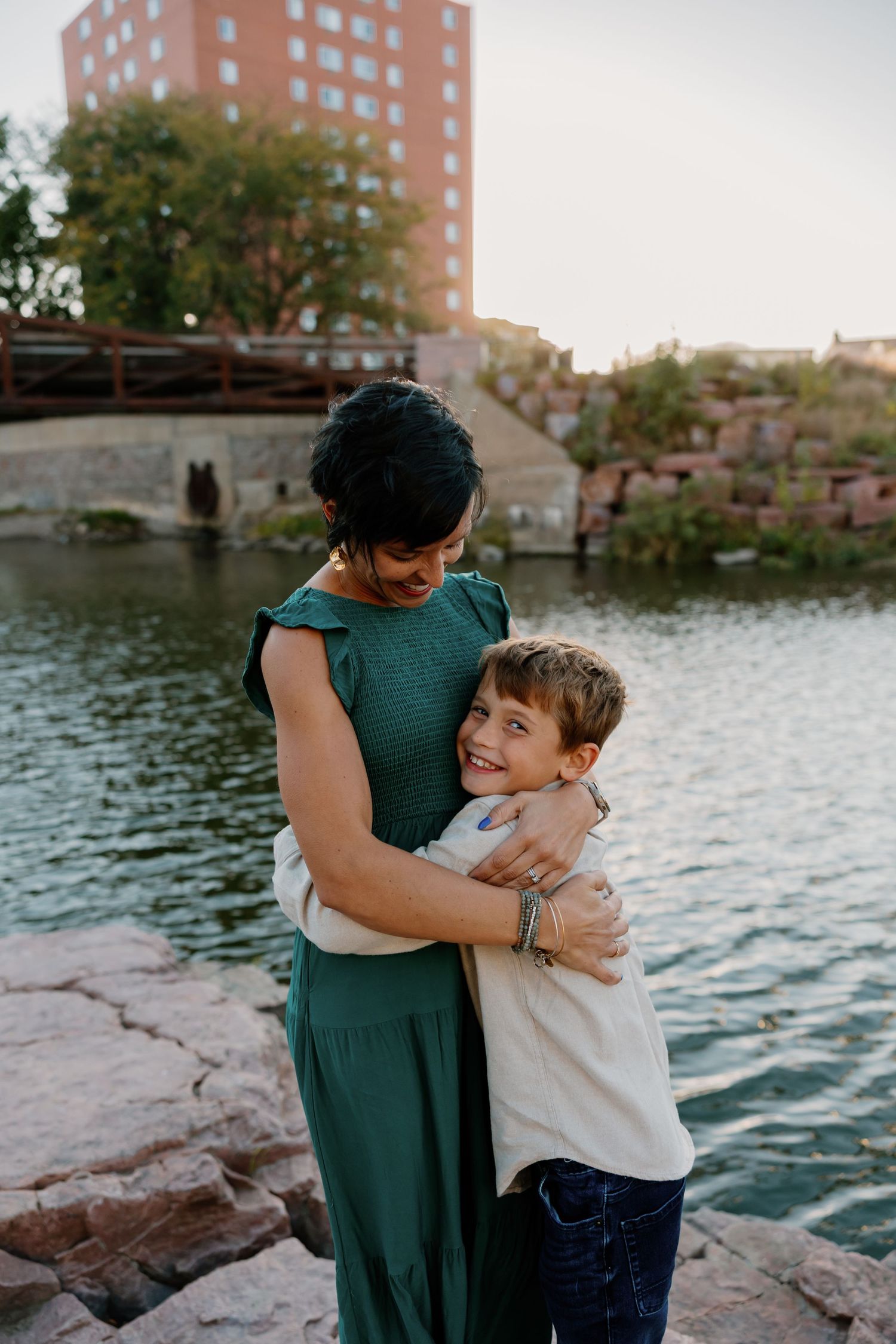 Mother and son share tender moments by a lakeside with brick buildings in the background during sunset. Riley Ro Photo Sioux Falls, SD