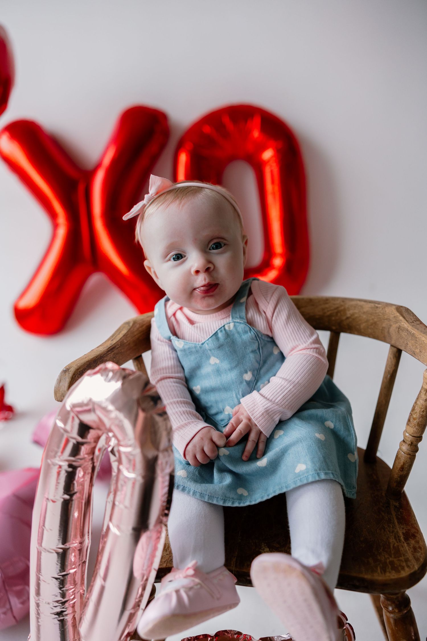 Small baby in light blue dress and pink tights sits on wooden chair with red balloon decorations in background.