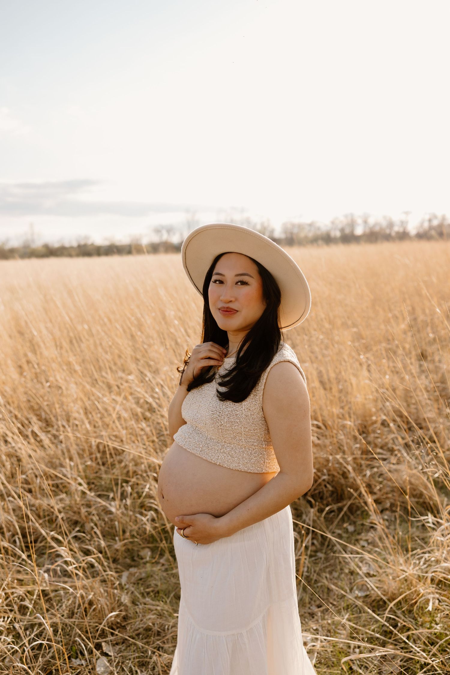 A pregnant woman in a white dress and wide-brimmed hat stands in a golden wheat field at sunset.