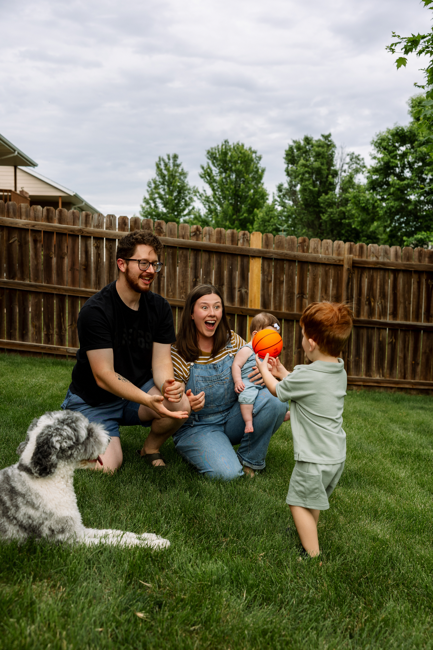 Family with two kids and a dog playing in a fenced backyard on a cloudy day, sharing joyful smiles.