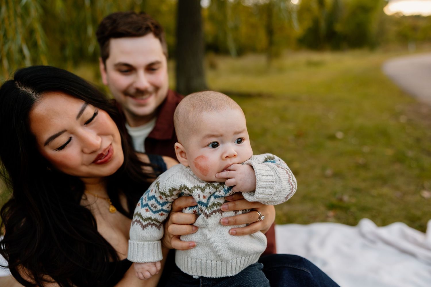 Parents sharing loving moments with their baby while relaxing on a white blanket in a park. Riley Ro Photo Sioux Falls, SD